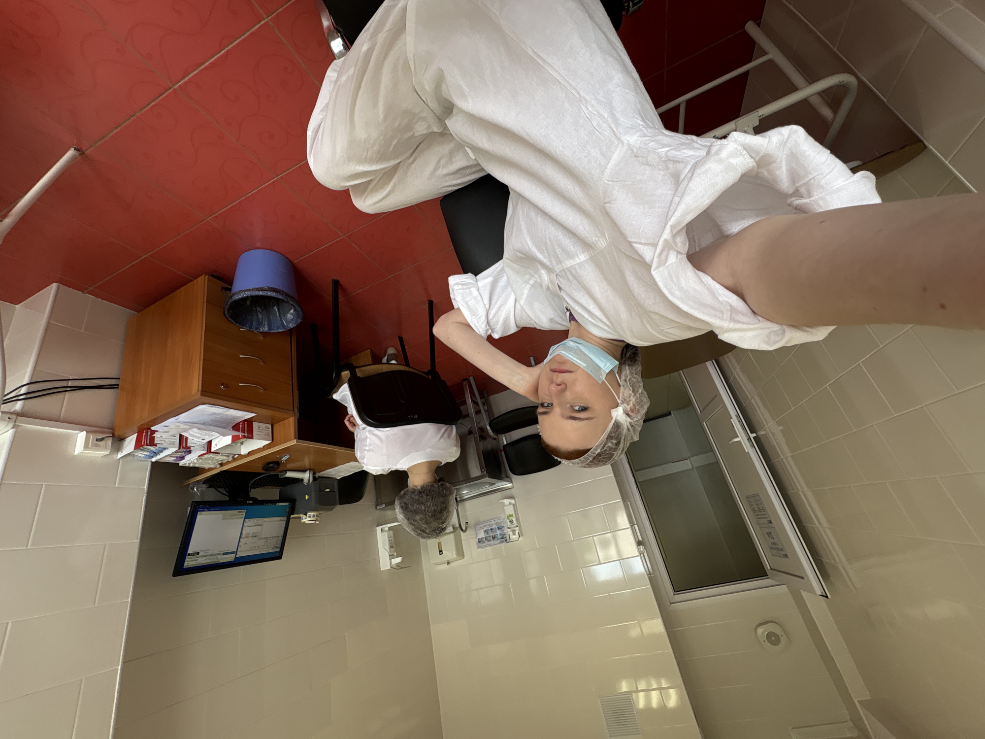 The image depicts a person in a white medical gown, a surgical cap, and a face mask, possibly in a medical facility. The person appears to be taking a selfie.
The environment includes tile walls, a computer monitor, a wooden cabinet with medical supplies, a blue trash bin, and a bed. The scene is indoors, likely within a clinic or hospital setting.
There is no information in the image that can confirm the location is in Saint Petersburg, Russia.