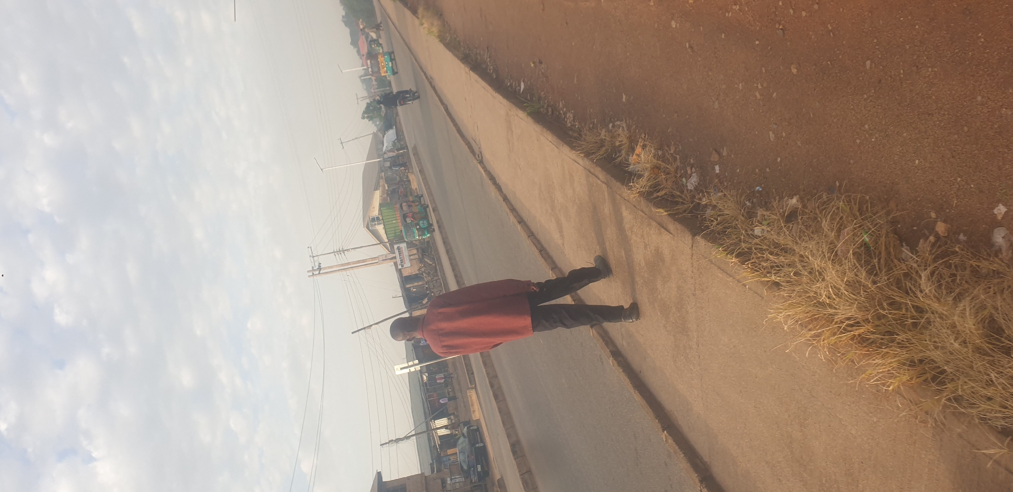A man wearing a rust-colored long-sleeved shirt and dark pants walks along a concrete sidewalk next to a paved road in Jos, Nigeria. The man is facing away from the camera, and his back is to the viewer. The road is lined with low-rise buildings, some appearing to be shops or market stalls. Several vehicles are visible on the road, including a black car, a green and yellow motorized tricycle, and a motorcycle. Power lines and utility poles are also present. The sky is overcast with scattered clouds. The sidewalk is bordered by dry grass and some debris on the right side. The overall scene depicts a typical street view in a commercial or residential area of Jos.