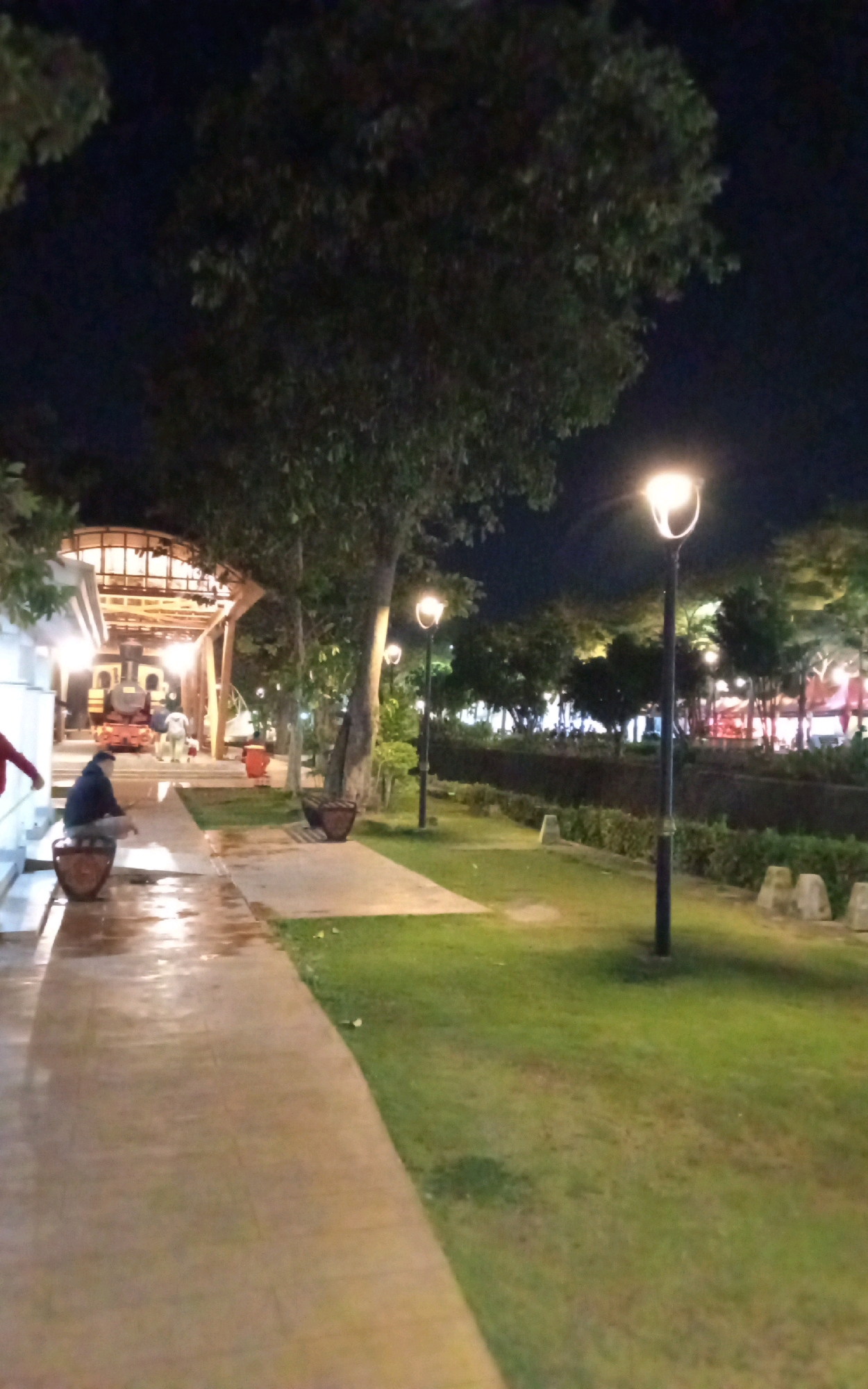 The image depicts an outdoor scene at night, likely a park or public area in the city of Ngasem, Indonesia. A paved walkway curves through the foreground, with a grassy area to the right. Several lampposts illuminate the scene. In the background, a building with warm interior lighting is visible, along with what appears to be a train or locomotive.
In the foreground, a person is seated on a planter or bench along the walkway, facing away from the camera. Their attire is dark. Another person, partially visible on the far left, is wearing red. The walkway appears to be wet, suggesting recent rain or cleaning. The overall atmosphere is calm and serene, illuminated by artificial light in the darkness.