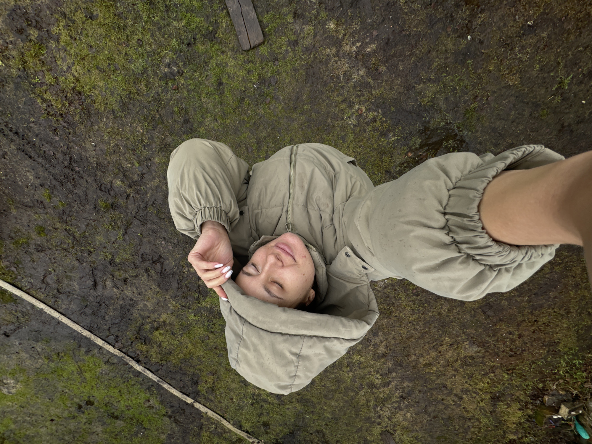 A high-angle shot shows a woman lying on her back on a muddy and grassy ground. She wears a light green puffy jacket with the hood up. Her eyes are closed and her hand is resting on her head. A partial view of a fair-skinned arm extends into the frame at the right. Some pieces of wood lay in the top center of the image, a hose in the bottom left of the image.