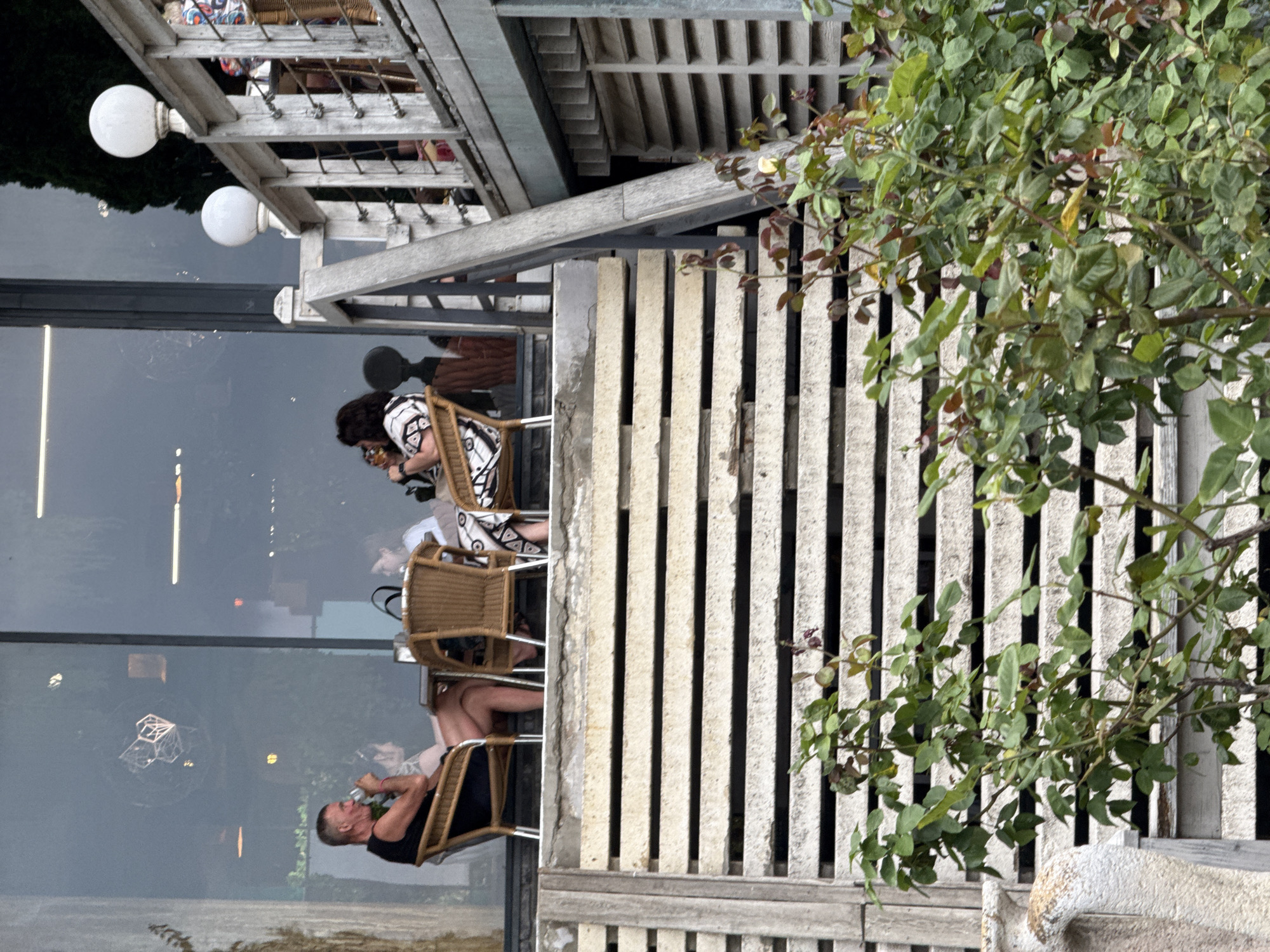 A group of four people are seated at tables on an elevated outdoor patio. The patio is accessed by a wooden staircase. The people appear to be dining or drinking. One man is wearing a black tank top and shorts, holding a drink. Two women are seated at the same table as the man, facing away from the camera. Another woman is seated at a separate table, facing the camera. She is wearing a black and white patterned top. The background shows a building with large windows reflecting the outdoors. There are two white globe lights mounted on a wooden railing to the right of the scene. Plants are visible in the foreground, obscuring part of the wooden staircase.