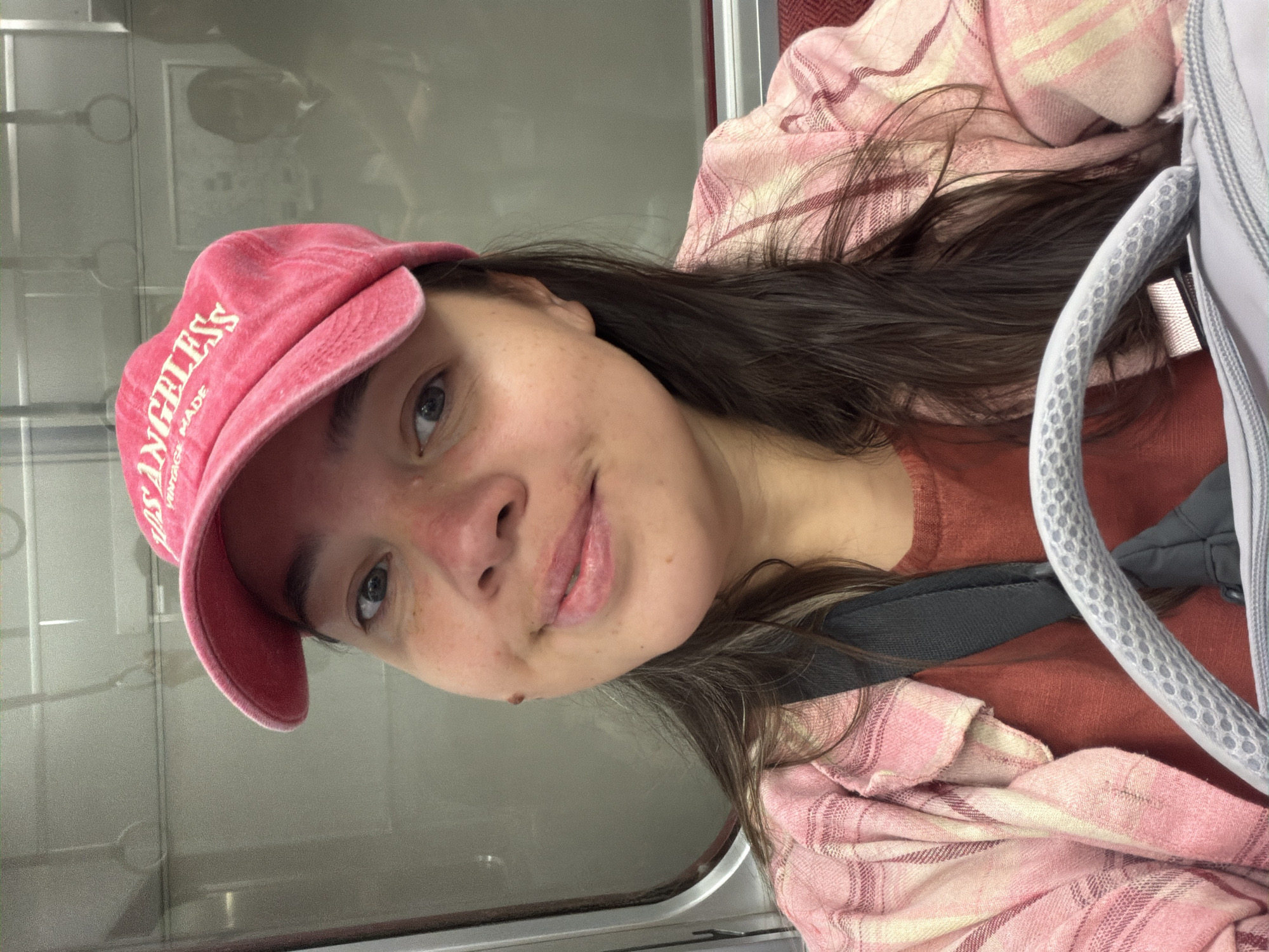 In a selfie taken indoors, a person with long, dark hair and a pink baseball cap with "LOS ANGELES" embroidered on it is looking at the camera. They are wearing a pink plaid shirt over a rust-colored t-shirt. The background appears to be the interior of a vehicle, possibly a train, with a window on the left and a headrest visible on the right. The person has freckles on their face and a faint smile.