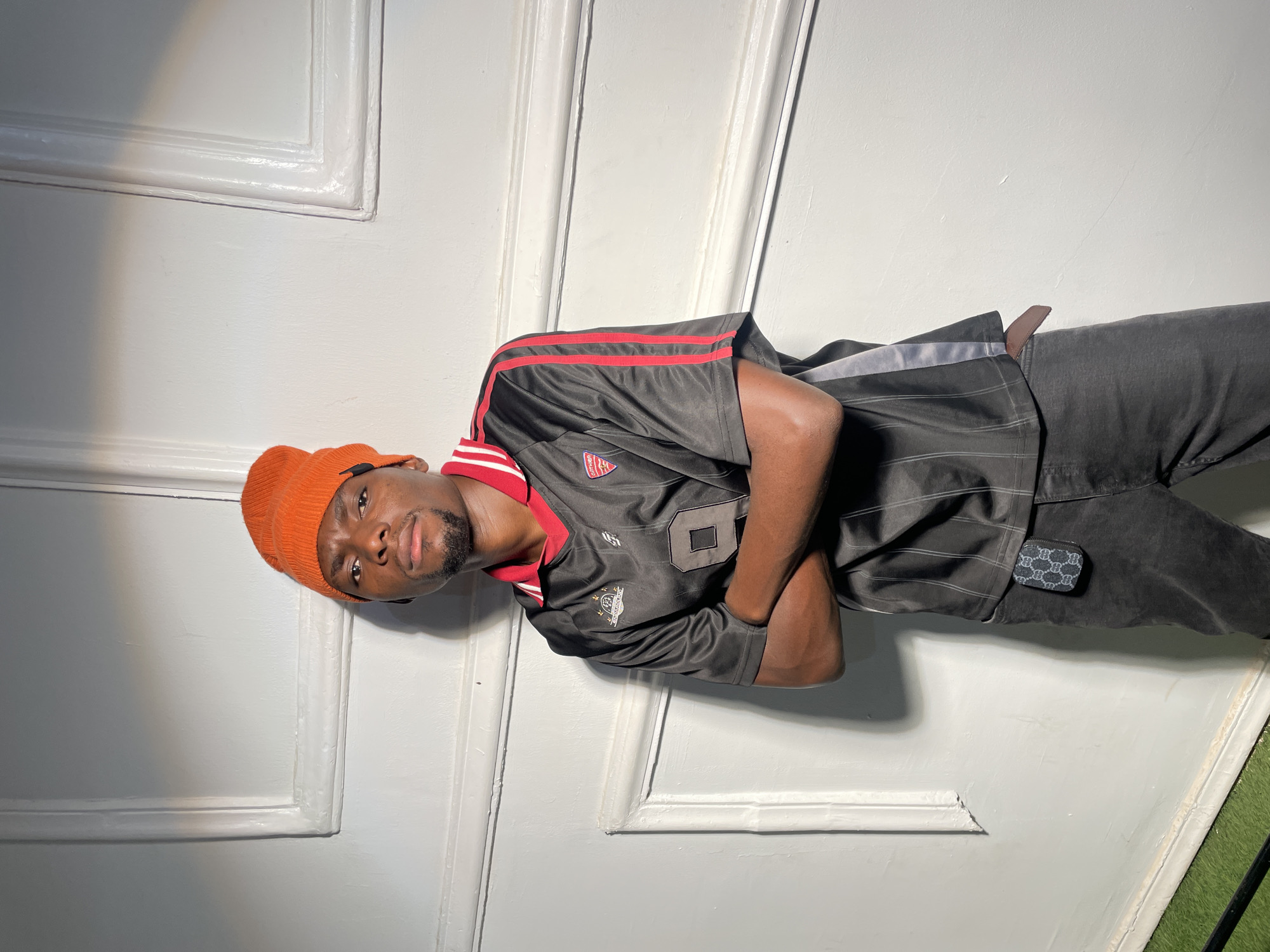 A dark-skinned man wearing an orange beanie, a black soccer jersey with red and white trim, and gray jeans is standing in front of a white paneled door. He has his arms crossed and is looking at the camera. The scene appears to be indoors. There is green artificial turf visible in the lower right corner of the image. The location is Barakin Akawo, Nigeria.