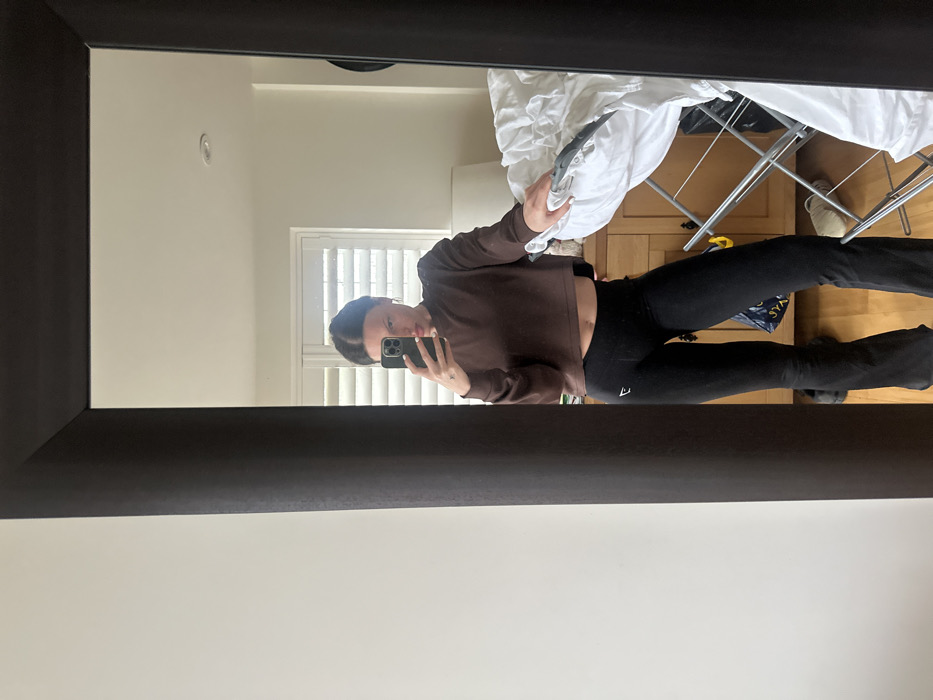 The scene depicts an individual taking a mirror selfie in an indoor residential setting, located within a room in Tunbridge Wells, United Kingdom. The subject, a female with dark hair styled away from her face, stands reflected in a large, dark-framed full-length mirror. She holds a dark-colored smartphone in her right hand at chest height, pointed towards the mirror to capture the image. A ring is visible on her left ring finger. She is dressed in a brown long-sleeved cropped top, which exposes her midriff, and black flared leggings featuring a white "Gymshark" logo on the upper left thigh.
The background visible in the mirror's reflection includes a light-colored wall with a window fitted with white louvered blinds. Below the window, a light wooden dresser or cabinet is present. To the right, a silver-colored collapsible clothes airer holds a substantial amount of white laundry, primarily crumpled fabric items, likely bed linens. A pair of light-colored athletic shoes are on the floor near the base of the airer, alongside a dark-colored bag. The floor appears to be light wood. The room is well-lit, suggesting natural light entry.