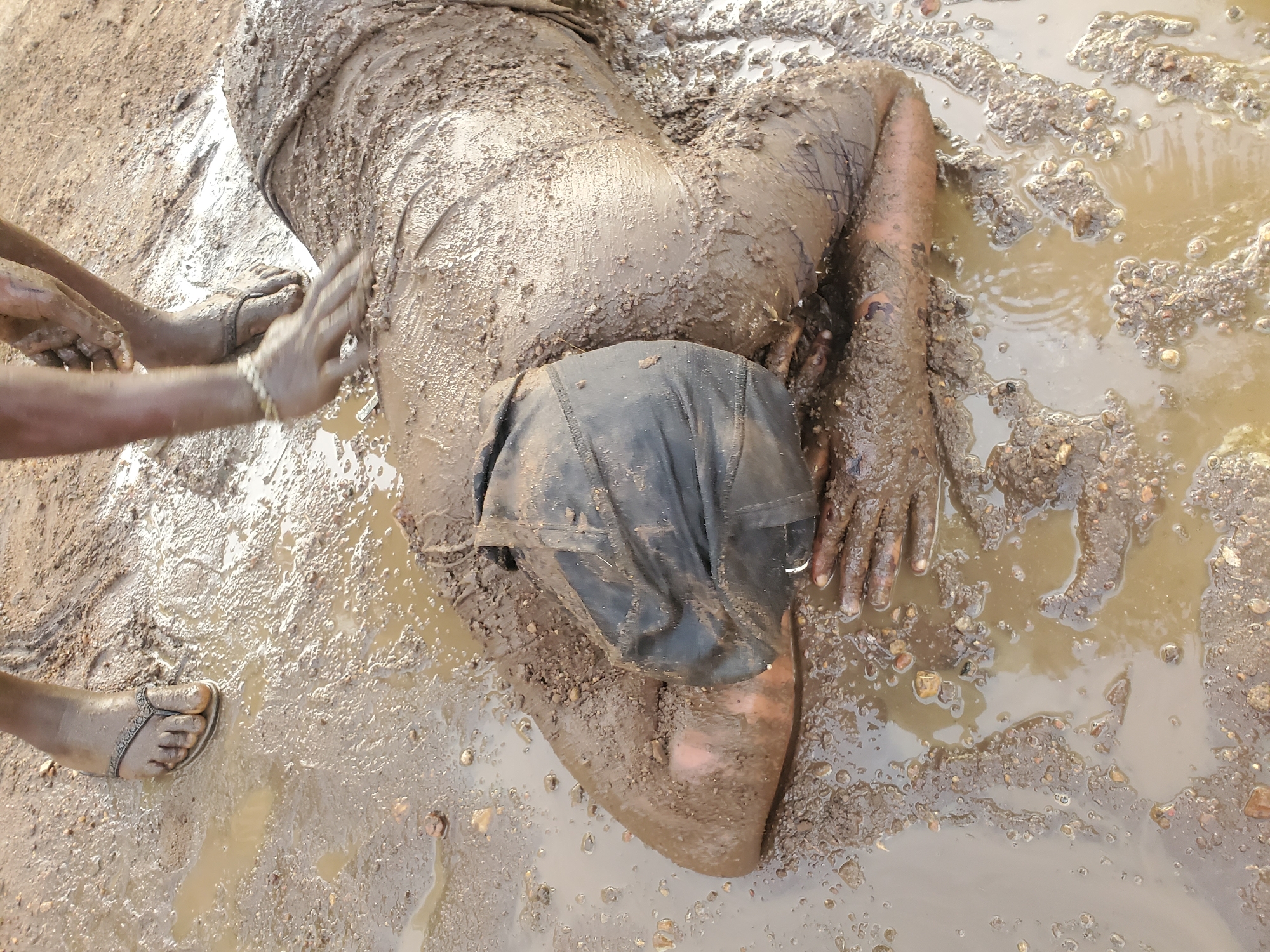 An overhead shot captures a scene set in a muddy, waterlogged environment, characteristic of Akwanga, Nigeria. The primary subject is a person lying prone, face down, almost entirely covered in thick, brown mud that adheres to their clothing and exposed skin. A dark piece of fabric or clothing covers the back of their head or upper back.
Interacting with this person are the hands and feet of a second individual, both also coated in mud. The second person's hands are positioned near the first person's lower back, seemingly either assisting them, applying mud, or cleaning them. One foot of the second person, wearing a simple sandal or flip-flop, is visible to the left.
The ground is a mix of wet mud, shallow puddles of brownish water, and scattered small stones or gravel. The water shows ripples in places, suggesting recent movement or drops. The general atmosphere suggests a very wet and possibly outdoor setting, likely after rain or in a consistently damp area. The lighting indicates it is daytime. No discernible text is visible in the image. The emotions or specific purpose of the activity are not clearly evident from the visual information alone.