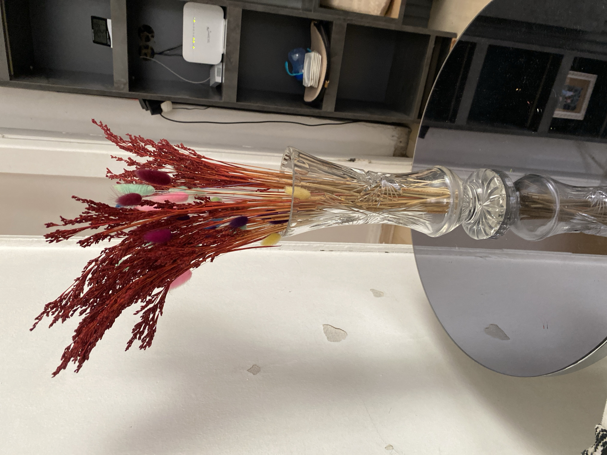A still life photograph captures a bouquet of dried red and multicolored decorative grasses in a clear, cut-glass vase. The vase sits on a dark, reflective circular table. Behind the table, a white wall with visible imperfections and patches of peeling paint is to the left, and a tall, dark shelving unit is to the right. The shelving unit contains a white router with illuminated indicator lights on the middle shelf, and a small digital clock-like device on the top shelf. The flooring is not visible. The setting appears to be an indoor residential space, potentially in Küçükçekmece, Turkey, given the provided location context. No people are present in the frame.