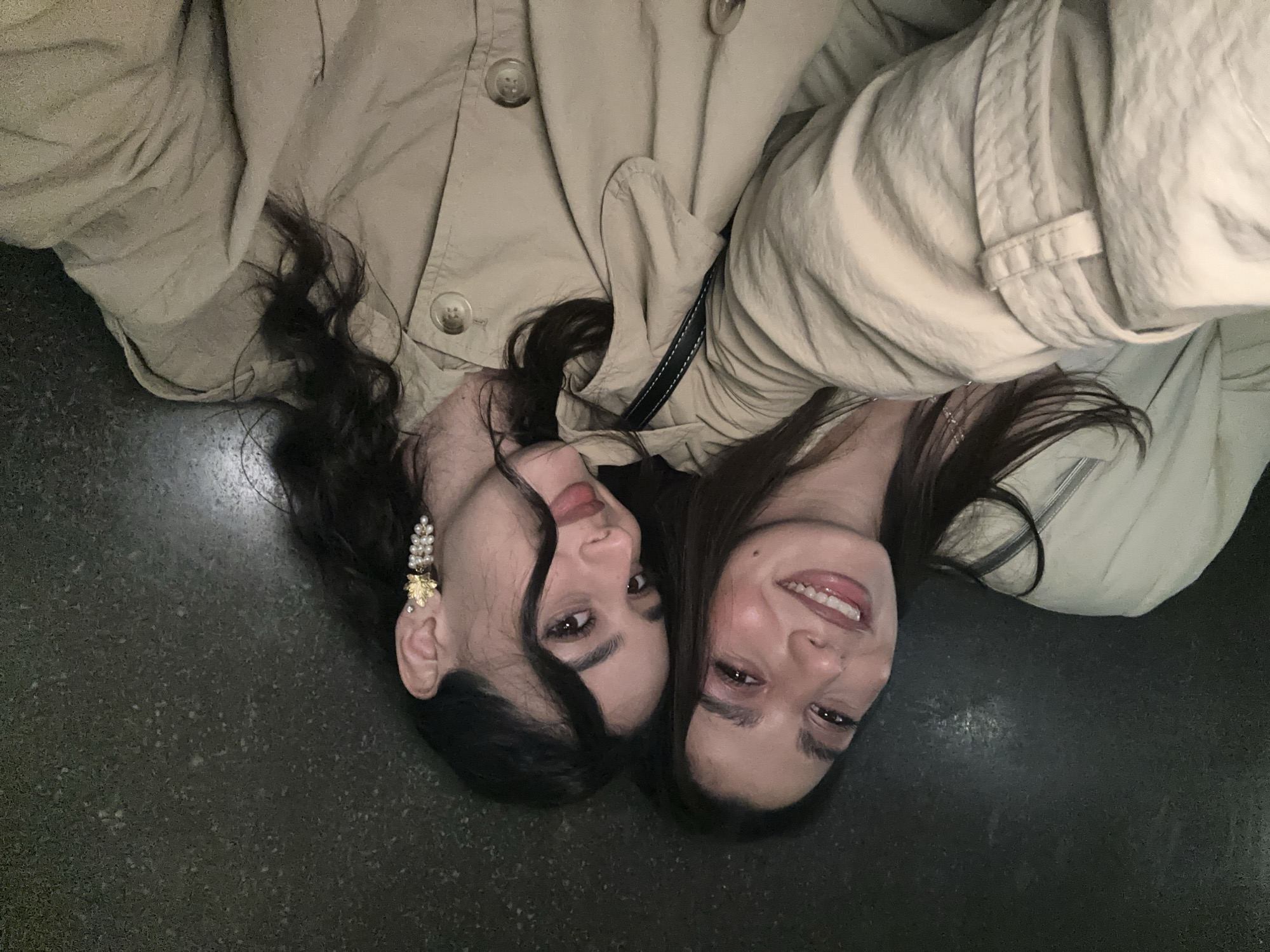 Two women are lying on a dark, possibly stone, surface. The woman on the left has dark, curly hair, an ornate pearl earring, and is wearing a light-colored trench coat. The woman on the right has straight, dark hair and is also wearing a light-colored trench coat. Their heads are close together, and they are both smiling. A black shoulder bag strap is visible between them. The surface appears to be a public space, possibly a plaza, in a city. It is impossible to definitively determine the location is Milan, Italy, from this image.