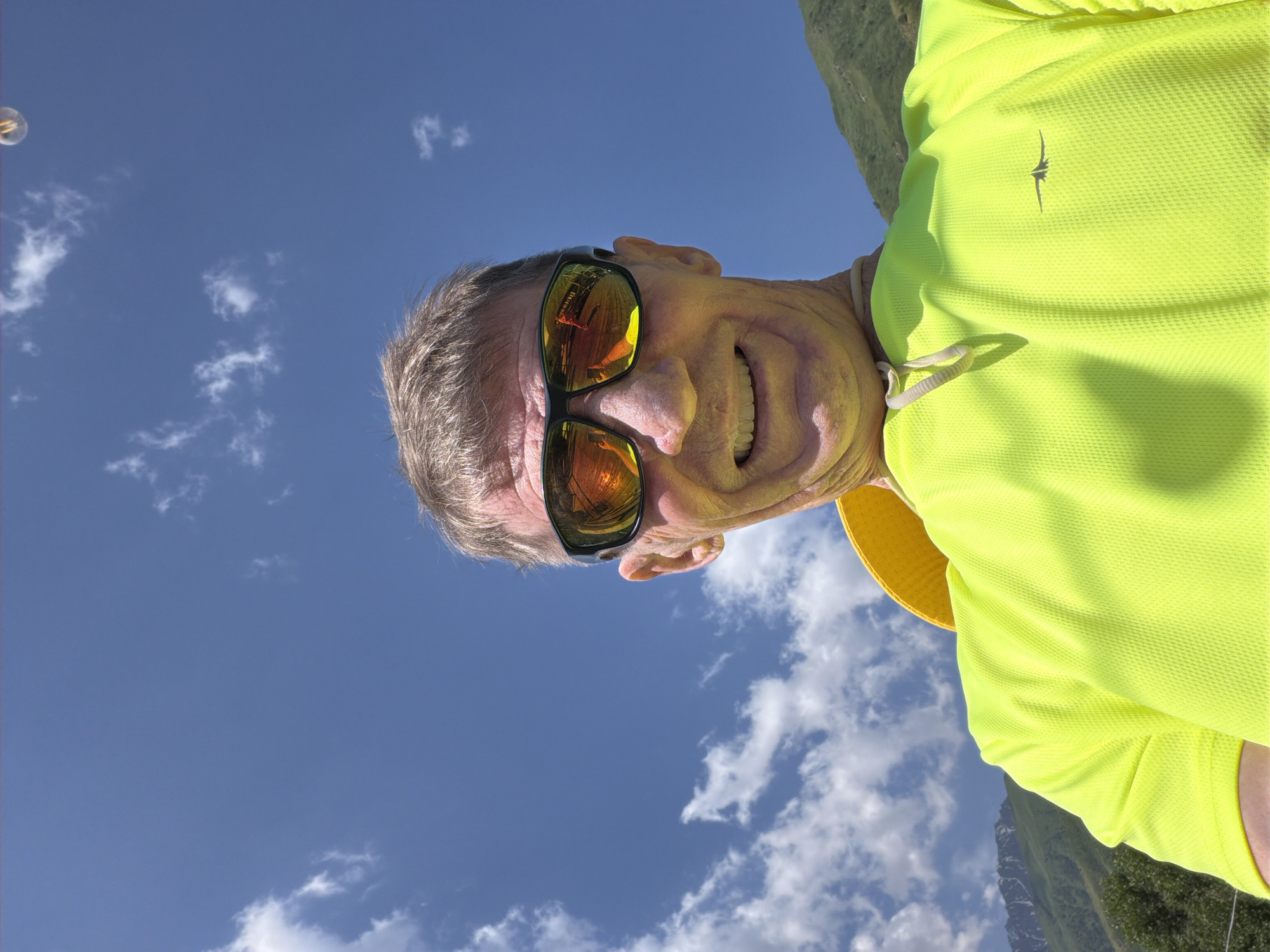 In a selfie taken from a low angle, a man with short, graying hair and a bright yellow shirt smiles broadly at the camera. He is wearing dark sunglasses with orange lenses that reflect a scene of trees, a building, and his own hand holding the camera. A yellow hat is visible around his neck. The background consists of a clear blue sky with scattered white clouds, and the green, mountainous terrain of a natural landscape. The scene is likely outdoors, given the clear sky and natural surroundings.