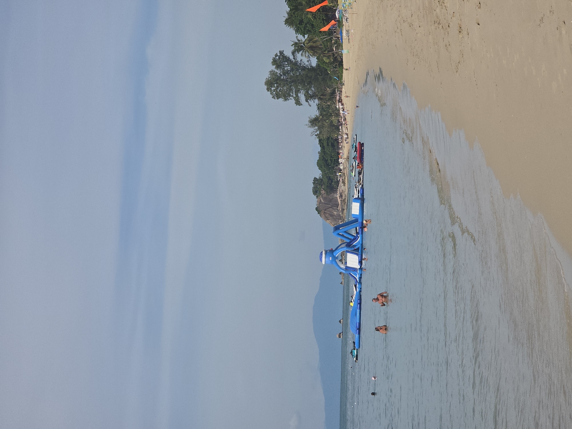 The image depicts a tropical beach scene with calm, clear blue water and a sandy shore. In the water, there is a large inflatable water park with slides and climbing structures, as well as several jet skis and a red inflatable boat. A few people are visible in the water, some swimming and others interacting with the inflatable park. The beach is lined with palm trees and other vegetation, and in the distance, mountains can be seen. The sky is partly cloudy with a light blue hue. There are orange flags planted on the beach. The overall atmosphere is one of leisure and recreation.