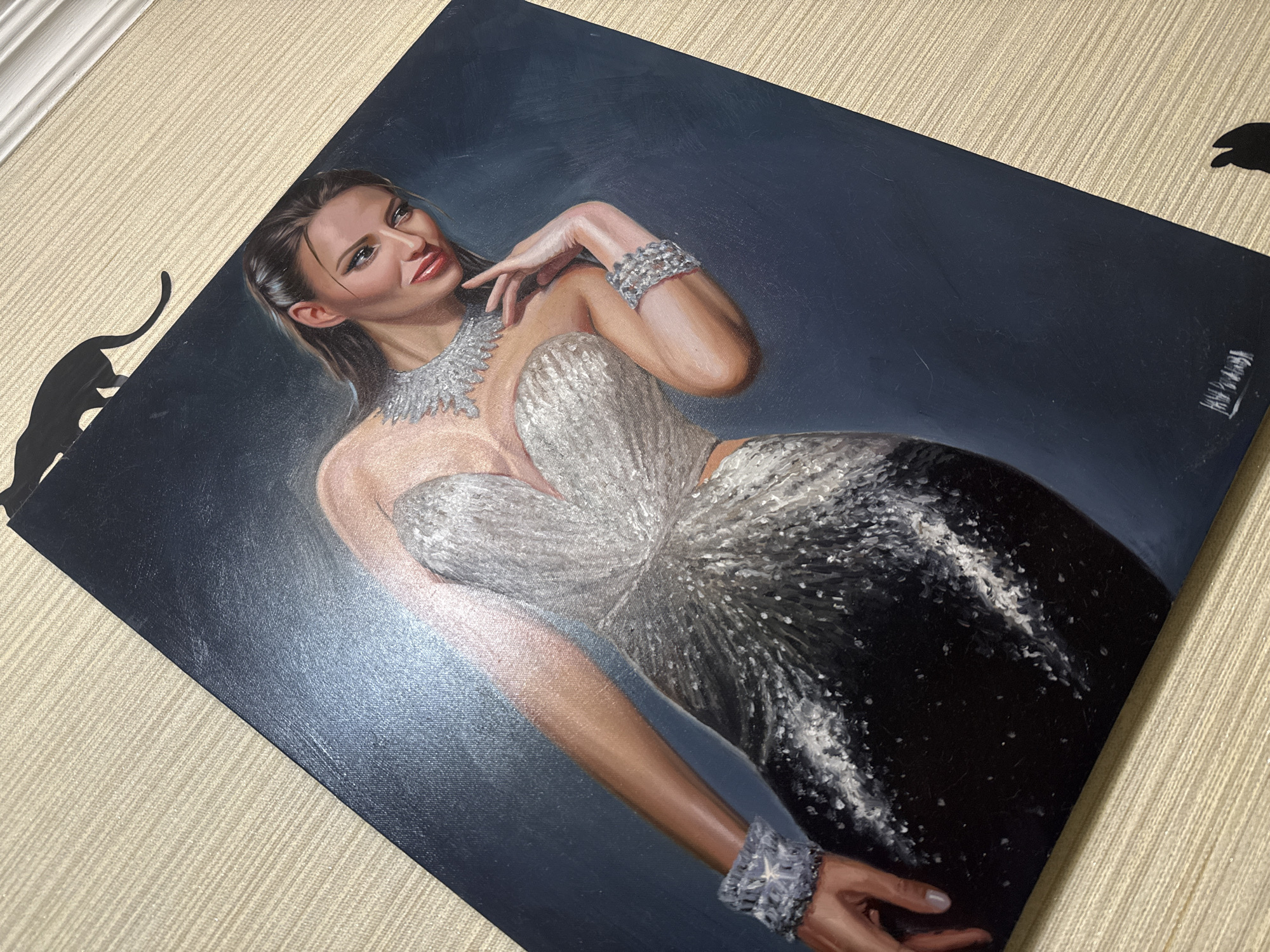The image is a high-angle shot of a painting on a textured beige surface. The painting depicts a woman in a shimmering silver and black gown with elaborate jewelry around her neck and wrists. She has dark hair styled back, and her gaze is directed to her right. Her right hand is near her face, with her fingers lightly touching her cheek. The background of the painting is a dark blue gradient. There are two black cat silhouettes positioned at the top and bottom right corners, partially obscuring the painting's edges. The painting is signed by the artist in white lettering at the bottom right. The beige surface appears to be a table or wall covering with a ribbed texture. No specific location in Simferopol, Ukraine, can be determined from the provided image.