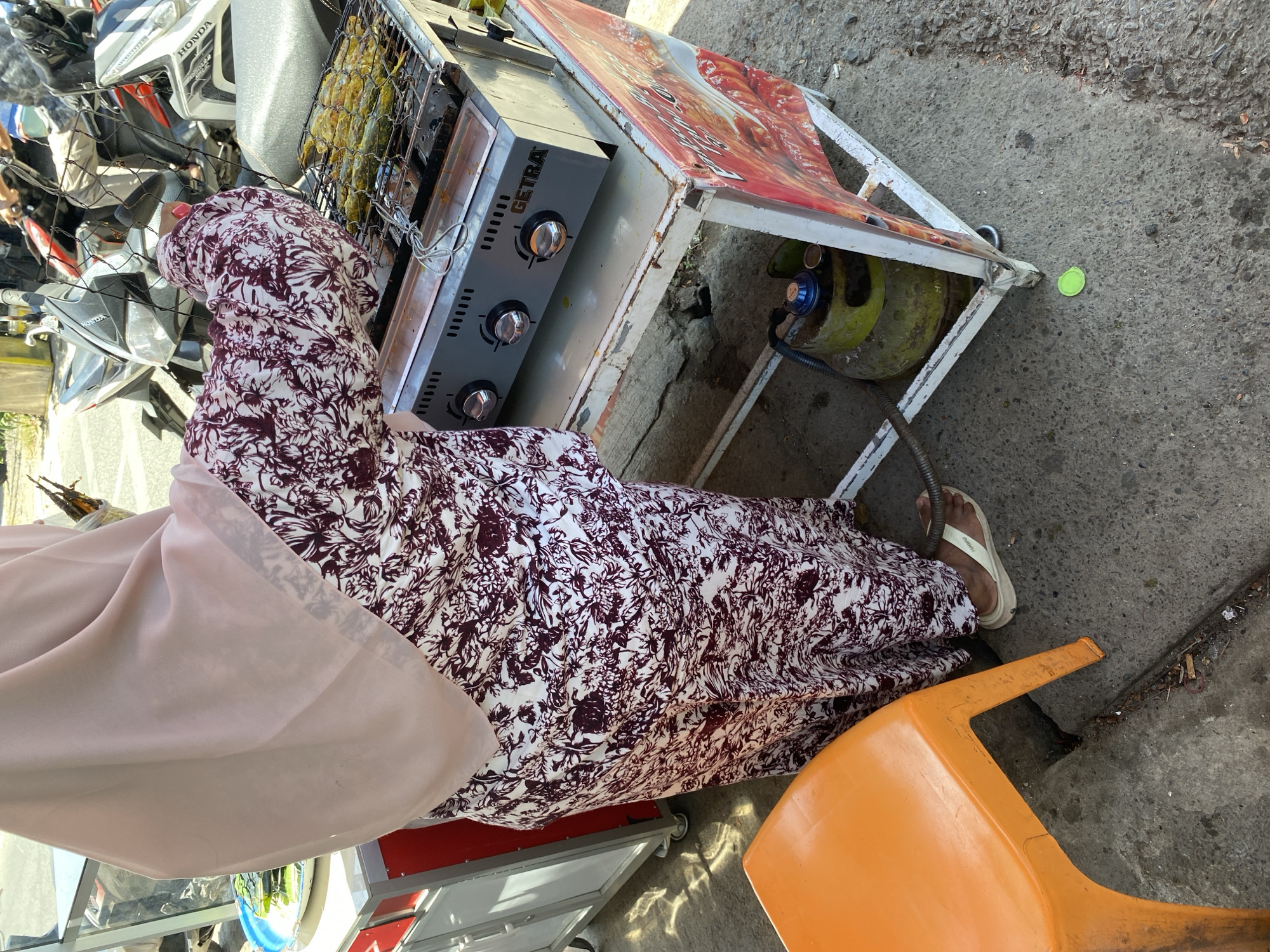A person wearing a pink hijab and a white and maroon floral dress is standing in front of a food stall in Bayu, Indonesia. The person is cooking fish on a gas grill with three burners, labeled "GETRA". The grill is placed on a metal cart with a red and white advertisement on the side. A green gas cylinder is connected to the grill. An orange plastic chair is visible in the foreground. In the background, motorcycles are parked. The scene appears to be outdoors, on a street or sidewalk.