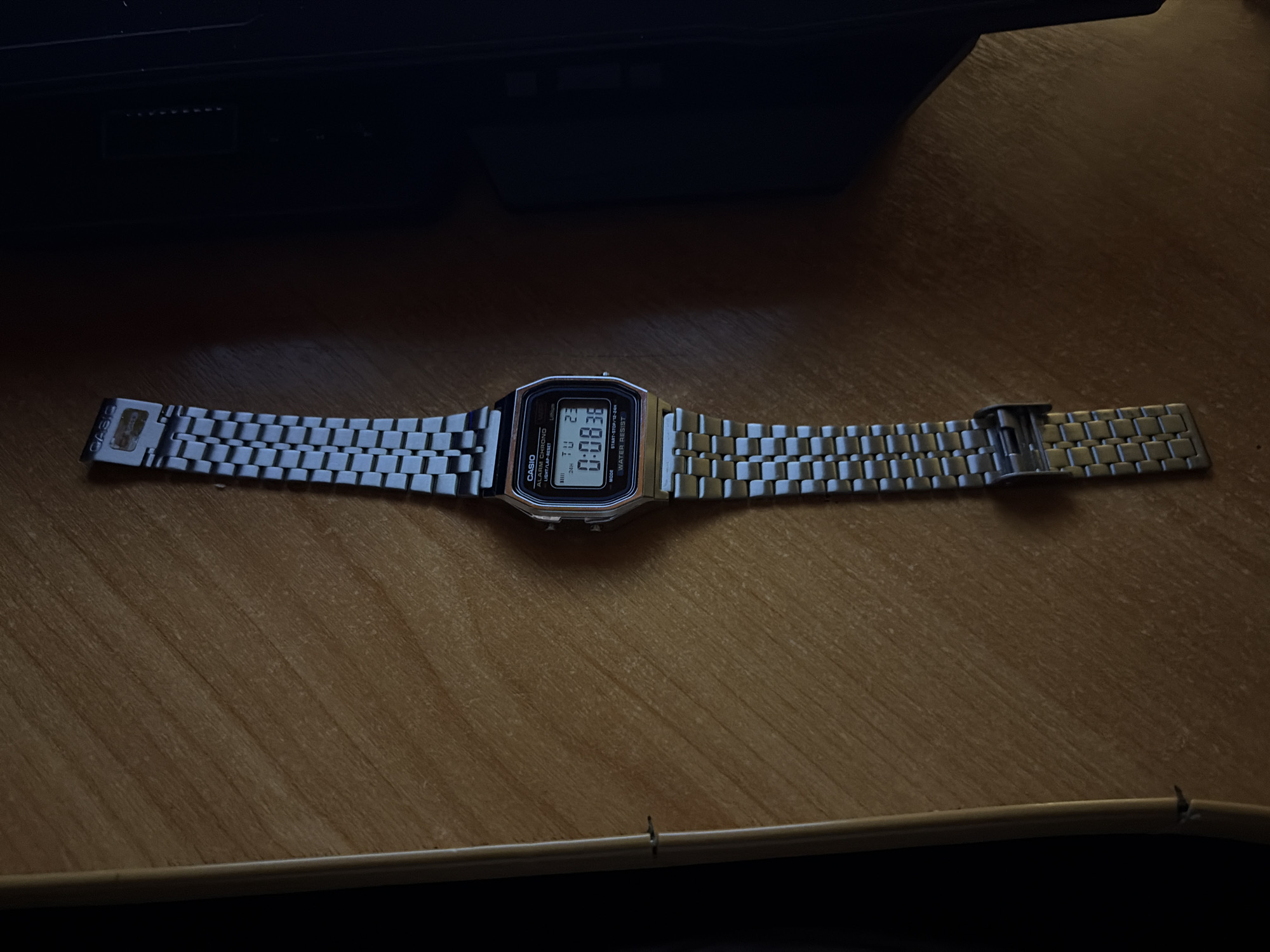 A silver Casio digital watch is lying on a wooden surface. The watch face displays "TU 23" at the top and "0:08'36" in larger numerals. Below the time, it reads "MODE" and "WATER RESIST". The watch has a metal link band. The wooden surface has a visible grain. The background is dark and out of focus. No specific location in Chistopol, Russia is identifiable from the image.