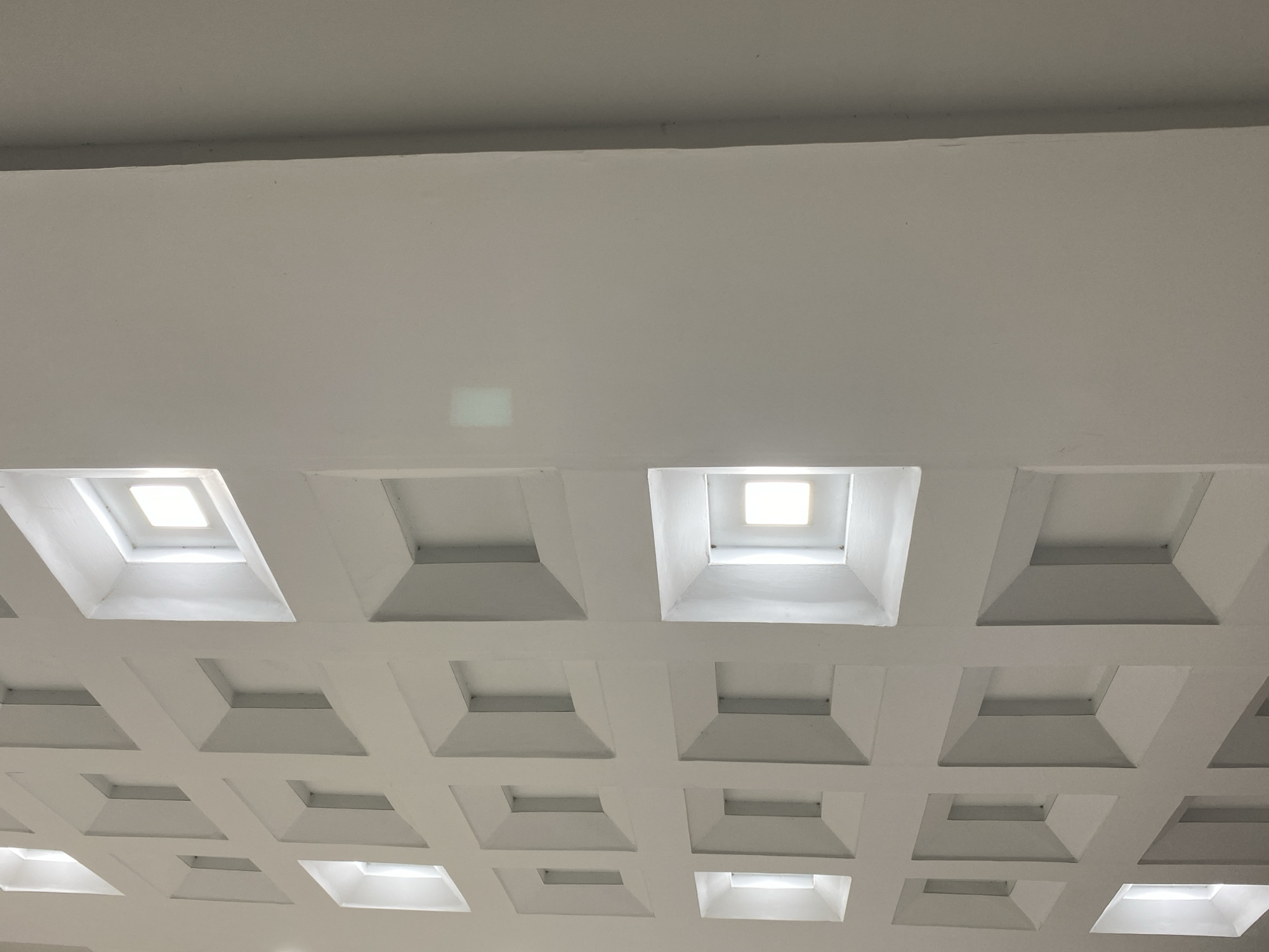 The image shows a white, coffered ceiling with square recesses. Some of these recesses contain light fixtures, emitting a soft glow. The ceiling has a geometric pattern created by the repeated square indentations. The lighting suggests an indoor environment, possibly a public building or a modern architectural space. The location in Banda Aceh, Indonesia, is not evident from the visual content of the image itself. There are no people, objects, actions, or interactions depicted.