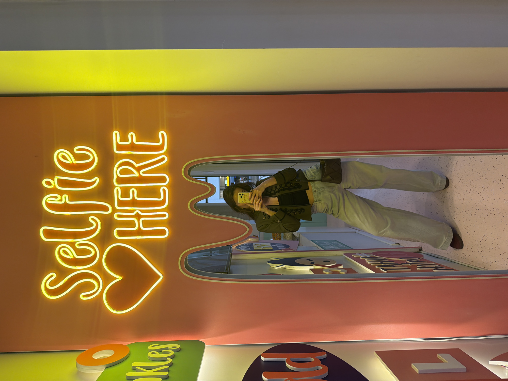 A person is captured taking a mirror selfie in a vibrant, brightly lit indoor setting, possibly in Belgrade, Serbia. The image is oriented vertically, showcasing the individual's reflection in a tall, arched mirror. The person, seen from the mid-torso downwards, is dressed in a dark jacket over a dark top, light-wash wide-legged jeans, and dark shoes. They carry a dark, possibly quilted, shoulder bag and hold a light-colored smartphone with a yellow case up to their face, obscuring their features as they take the picture.
The mirror is embedded in a prominent coral-pink wall. To the left of the mirror, a glowing yellow neon sign spells out "SELFIE HERE" in a stylized font, with a heart shape positioned beneath the word "HERE". Above the pink wall, a bright yellow horizontal band runs across, topped by a grey ceiling. The floor, visible both within the reflection and in the foreground, is light-colored with subtle speckles. At the very bottom of the image, below the pink wall, are decorative elements featuring colorful panels (green and purple) with large, cutout letters; one segment partially reads "kies". The scene suggests a playful and modern environment, likely a designated photo opportunity spot within a commercial or entertainment venue.