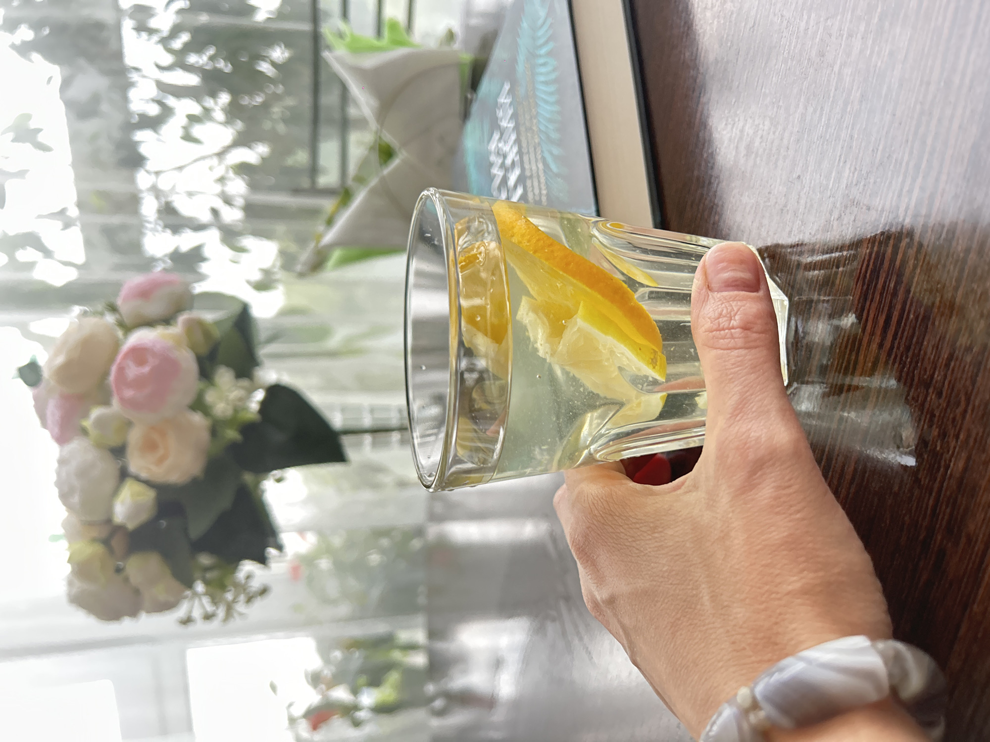 A close-up shot focuses on a hand holding a glass of water with lemon slices. The hand, adorned with a bracelet made of grey and white beads, is positioned in the foreground on a dark wooden table. Behind the glass, a book with a blue cover and a feathered design is visible, along with a tissue holder and a vase of flowers in the background. The scene appears to be indoors, possibly in a cafe or home setting, with natural light filtering in from a window. The location is not Kemerovo, Russia.