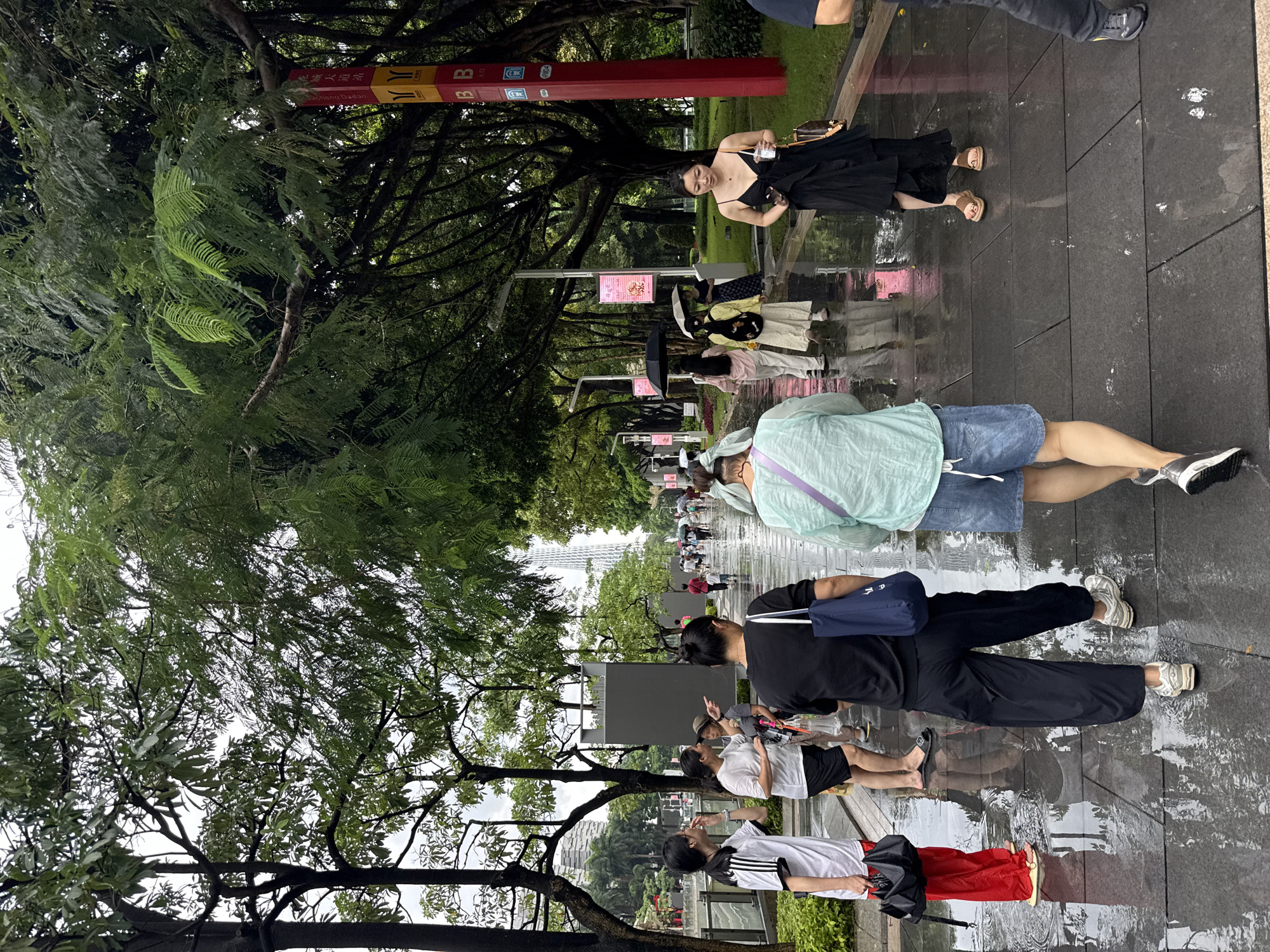 The image depicts a street scene in Tianhe District, China, following a rainfall. The ground is wet and reflective, indicating recent precipitation. Several individuals are walking along a paved pathway lined with trees and lush greenery. The architecture in the background suggests an urban environment, with tall buildings visible. Some people are holding umbrellas, and others are dressed in casual attire, including t-shirts, pants, and shorts. There are also some decorative elements like a red and yellow signpost with Chinese characters. The overall atmosphere appears to be one of a typical day in a city park or a pedestrianized street.