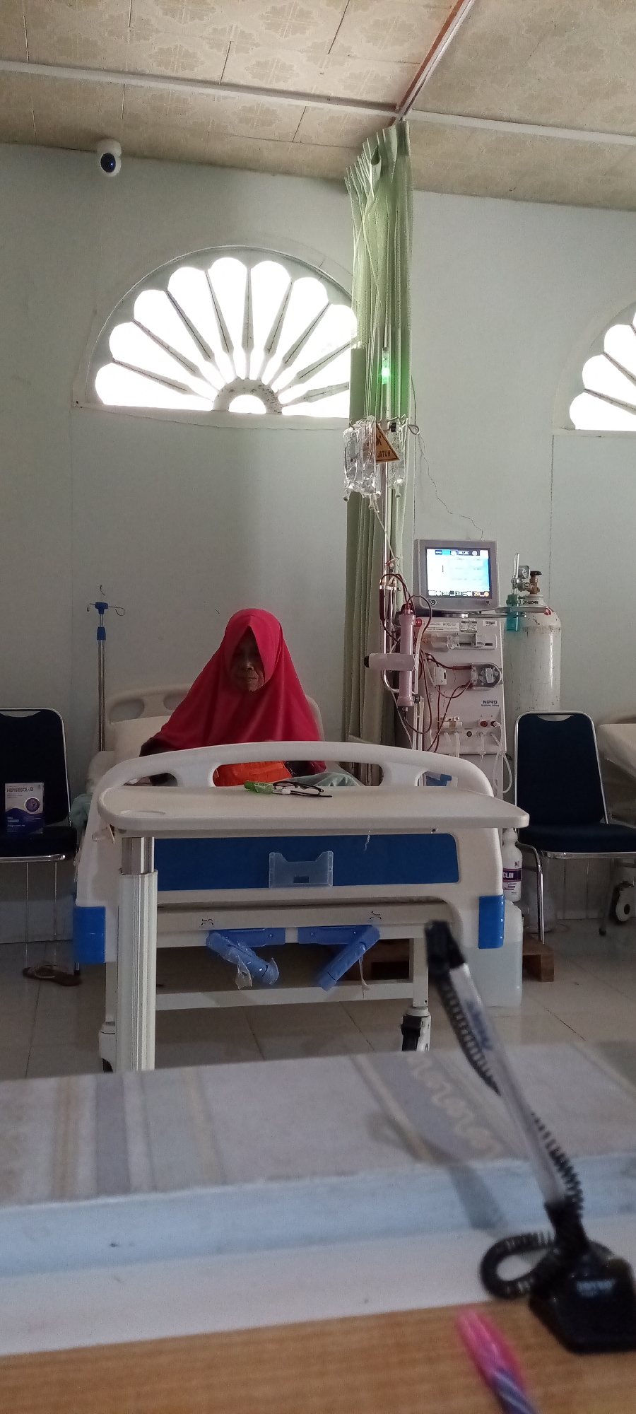 An elderly individual, likely a woman, wearing a bright red hijab, sits in a hospital bed in what appears to be a medical facility, possibly a dialysis unit, in Lhokseumawe, Indonesia. She is positioned centrally in the frame, looking towards the viewer with a calm expression.
To her right is an elaborate Nipro brand dialysis machine with a monitor displaying data and the Indonesian words "SELAMAT DATANG" (Welcome). An IV stand with two fluid bags hangs above the machine; one of the hangers has a label with the text "JATUH" (Fall/Dropping). Next to the dialysis machine is a gas cylinder. To the left of the bed, another IV pole stands by a dark chair, which holds a box labeled "NEPRESOL-D." A similar dark chair is placed to the right of the dialysis machine, and a white bottle with partially visible text "CLIN" is on the floor nearby.
The room is brightly lit by natural light streaming through two large, arched windows with decorative radial spokes, indicating it is daytime. Light green medical curtains are partially drawn near the medical equipment. The ceiling features square tiles, and a white security camera is mounted in the top-left corner of the room. The floor is tiled. In the immediate foreground, a wooden surface, possibly a desk or counter, is visible, with a black coiled pen and a pink/purple pen resting on it. Faint text, "LeeLoo," can be seen on the desk surface. The overall scene depicts a patient undergoing or awaiting medical treatment in a clinical setting.