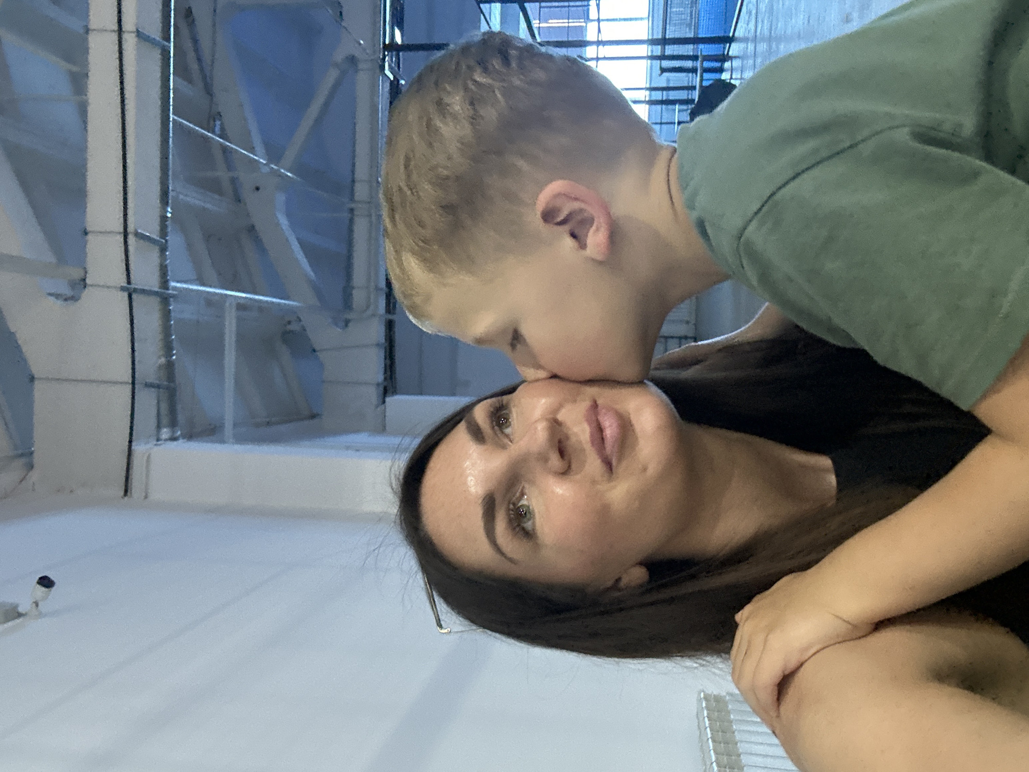 The media file is a photograph taken indoors. The primary subjects are a young boy and a woman. The boy, with blond hair, is leaning in to kiss the woman on the cheek. The woman, with dark hair, is looking towards the camera with a gentle expression. She is wearing glasses. The background shows industrial-looking metal structures and a white wall. There is a security camera visible on the white floor. The lighting suggests it is daytime. The image has a warm, affectionate tone. There is no visible text in the image.