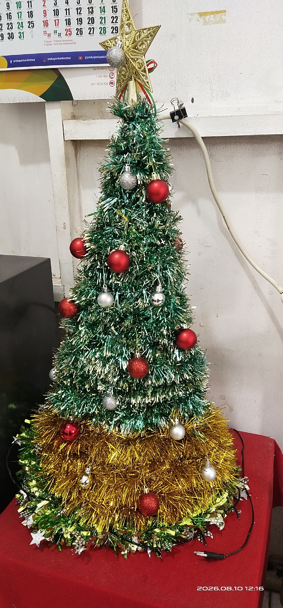 A tinsel Christmas tree, approximately two feet tall, stands on a red tablecloth. The tree is decorated with red and silver ball ornaments. A gold star ornament tops the tree, with green, white, and red ribbon accents. At the base of the tree is a large amount of gold tinsel. String lights with star-shaped ornaments are visible around the base. In the background, a calendar displays the month of August and the year 2023. A white wall with some discoloration is behind the tree, and a light-colored electrical cord runs from the top right of the frame down towards the tree. A black binder clip is attached to the wall, holding the cord in place. The scene is indoors, likely in a domestic or office setting. The timestamp on the image indicates it was taken on August 10, 2026, at 12:16 PM, which contradicts the calendar in the background.