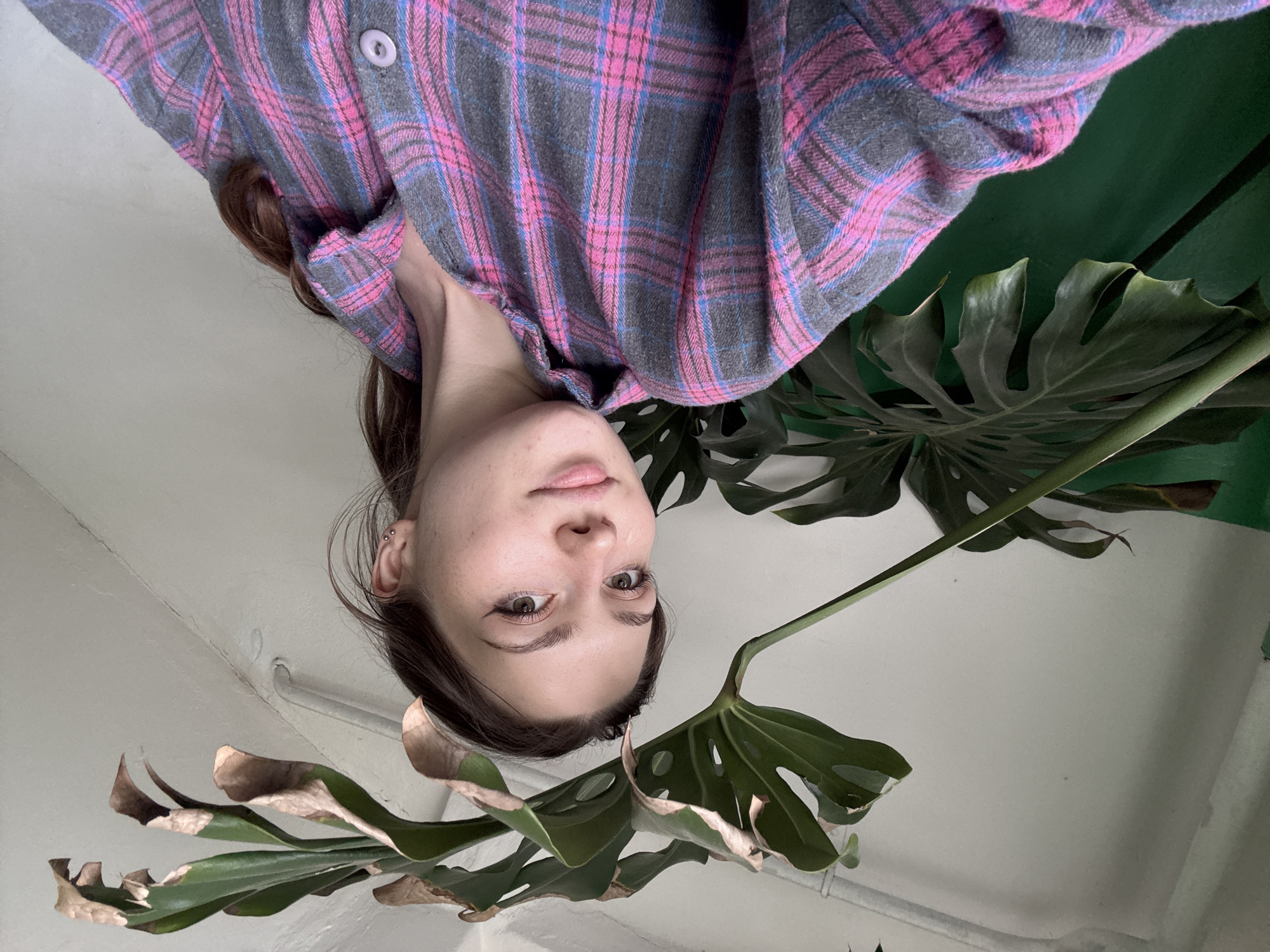 A young woman with brown hair, styled in a ponytail, is looking at the camera in a close-up shot. She is wearing a gray and pink plaid shirt. She has light-colored skin and is wearing subtle eye makeup, including winged eyeliner. She is also wearing small stud earrings. Behind her, there is a large green Monstera plant with several large leaves, some of which have brown, dried edges. The background is a plain, light-colored wall, and a metal pipe runs horizontally across the upper part of the image. The lighting suggests an indoor environment. The image does not provide any specific information about the location in Kumyonki, Russia.