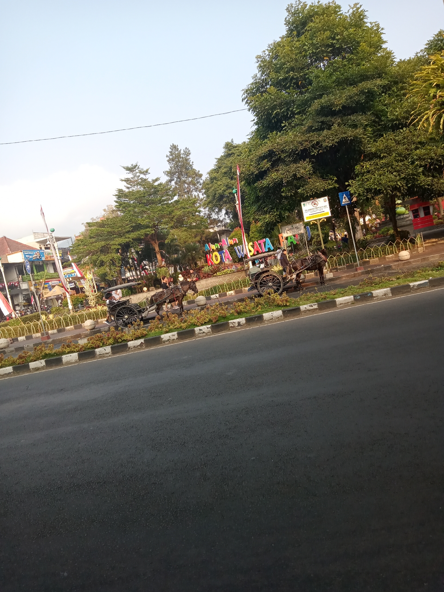 In Batu, Indonesia, a horse-drawn carriage carrying two people travels along a paved road beside a median strip. The median strip is adorned with plants and a decorative fence, with a large colorful sign displaying "KOTA WISATA BATU" (Batu Tourist City) behind it. Trees line the background, and various buildings and flags are visible. The sky is clear and bright.