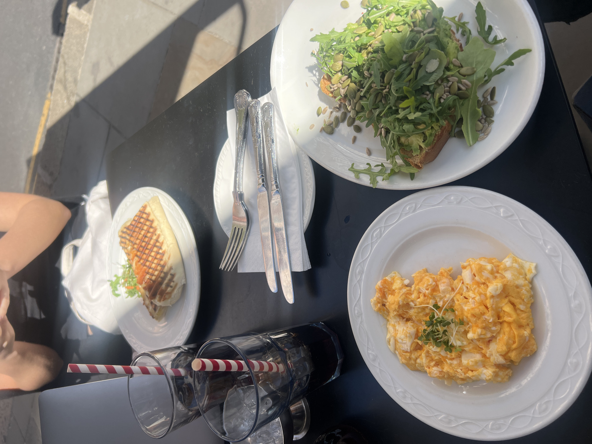 The image presents an overhead view of an outdoor dining table set for breakfast or brunch on a sunny day. Strong natural sunlight casts distinct, sharp shadows across the dark tabletop and the adjacent pavement, suggesting a morning or early afternoon setting.
Three white ceramic plates, each featuring a subtle embossed decorative rim, are arranged on the table. In the top right corner, a plate holds a slice of toast, generously topped with mashed avocado, a substantial amount of arugula or rocket leaves, and a sprinkle of pumpkin and sunflower seeds. To the bottom right, a second plate contains a large serving of fluffy scrambled eggs, garnished with a small cluster of fresh microgreens. On the left side, the third plate features a grilled sandwich, possibly a panini, with clear grill marks on its surface, accompanied by a small portion of salad leaves.
In the center of the table, a set of ornate silver-colored cutlery, consisting of a fork and two knives, is neatly placed on a white napkin, which rests on a small white side plate. Two clear drinking glasses, each fitted with a red and white striped paper straw, are positioned in the foreground.
A person's arm and leg are partially visible on the left side of the frame, indicating someone is seated at the table, about to or in the process of enjoying the meal. The visible pavement or cobblestones next to the table reinforce the outdoor, possibly street-side or patio, setting. The scene conveys a relaxed al fresco dining experience. No discernible text is visible on any of the items.