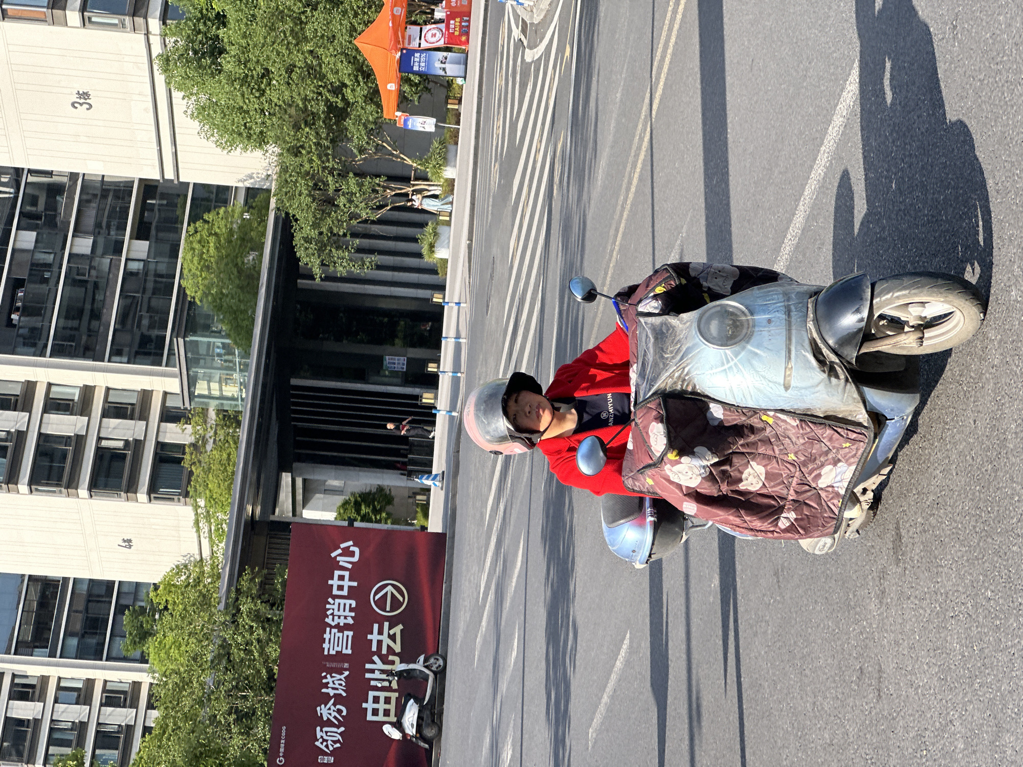 This bright and sunny image, taken in Jiangjin, China, depicts a street scene with a person riding an electric scooter. The long, distinct shadows on the asphalt suggest either morning or late afternoon.
In the foreground, a person wearing a helmet and a red jacket, with "ANZHIYUN" visible on it, is seated on a light-colored electric scooter. The scooter has a brown, quilted cover with cartoon patterns draped over its front section.
To the left of the rider, tall, modern buildings with multiple glass windows line the street. One building is marked with the characters "3栋" (Building 3). Green trees are visible both in the foreground and between the buildings, adding natural elements to the urban landscape.
A large red banner stands prominently on the left side of the frame, displaying Chinese characters that read "领秀城 营销中心 由此去" (Leading City Marketing Center, Go this way), accompanied by a right-pointing arrow. Below this, "G 中国绿发 CGDG" (China Green Development Group) is visible.
In the background, an orange canopy tent and several smaller signs are present. Text on these signs includes phrases like "国补至高 立省15%" (National subsidy up to 15% saving), "打话费" (Call charges), and "AI手机" (AI phone). The street itself is paved with asphalt and features clear white dashed and solid line markings. The scene captures a vibrant, modern city environment.