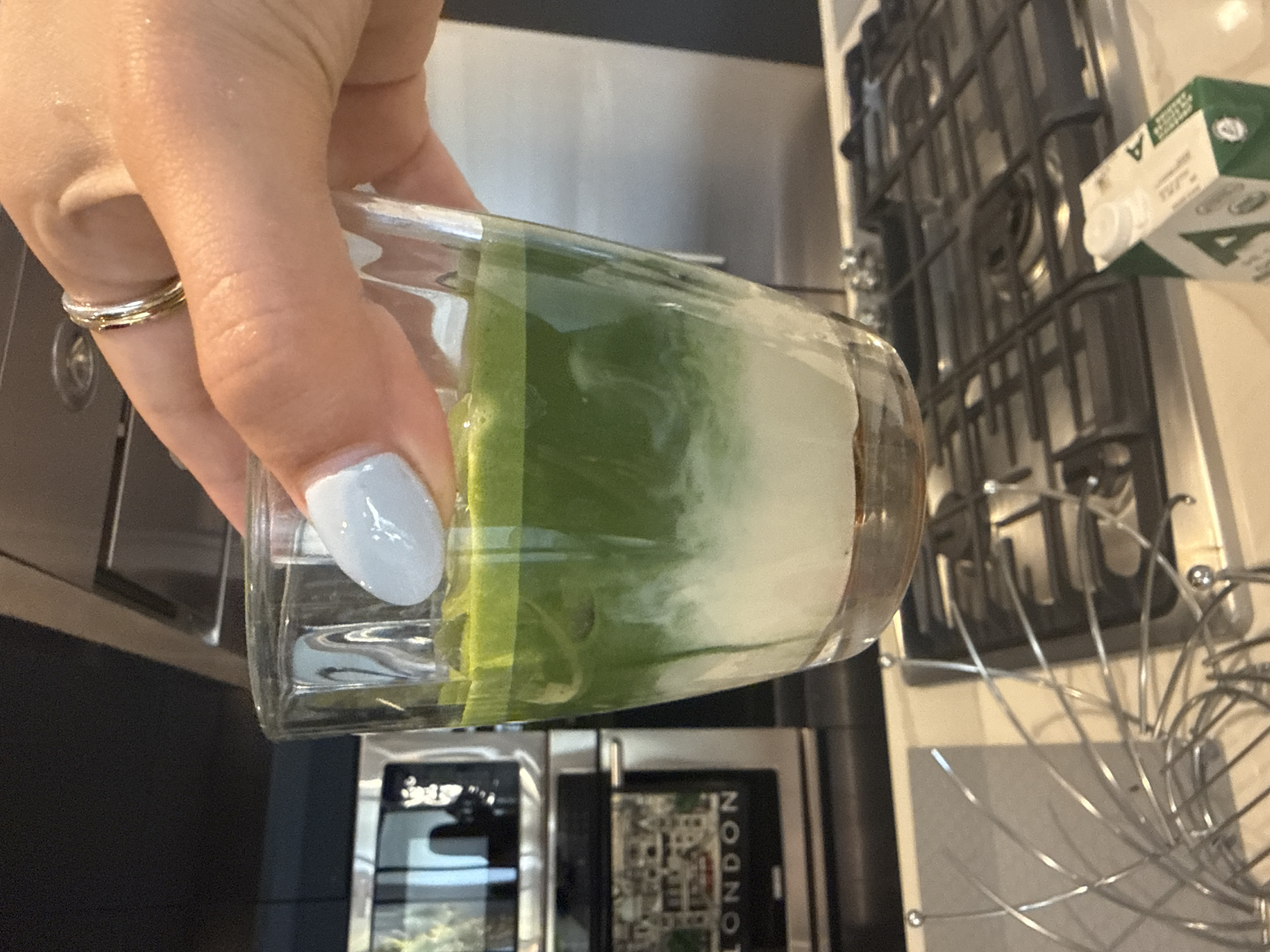 A hand with light blue nail polish and wearing two rings holds a layered drink in a clear glass. The drink has a brown layer at the bottom, a white layer in the middle, and a green layer on top. The background includes a coffee maker with "London" printed on its panel, a stove top, and a carton of milk. The image was not taken in San Bernardino Tlaxcalancingo, Mexico.
