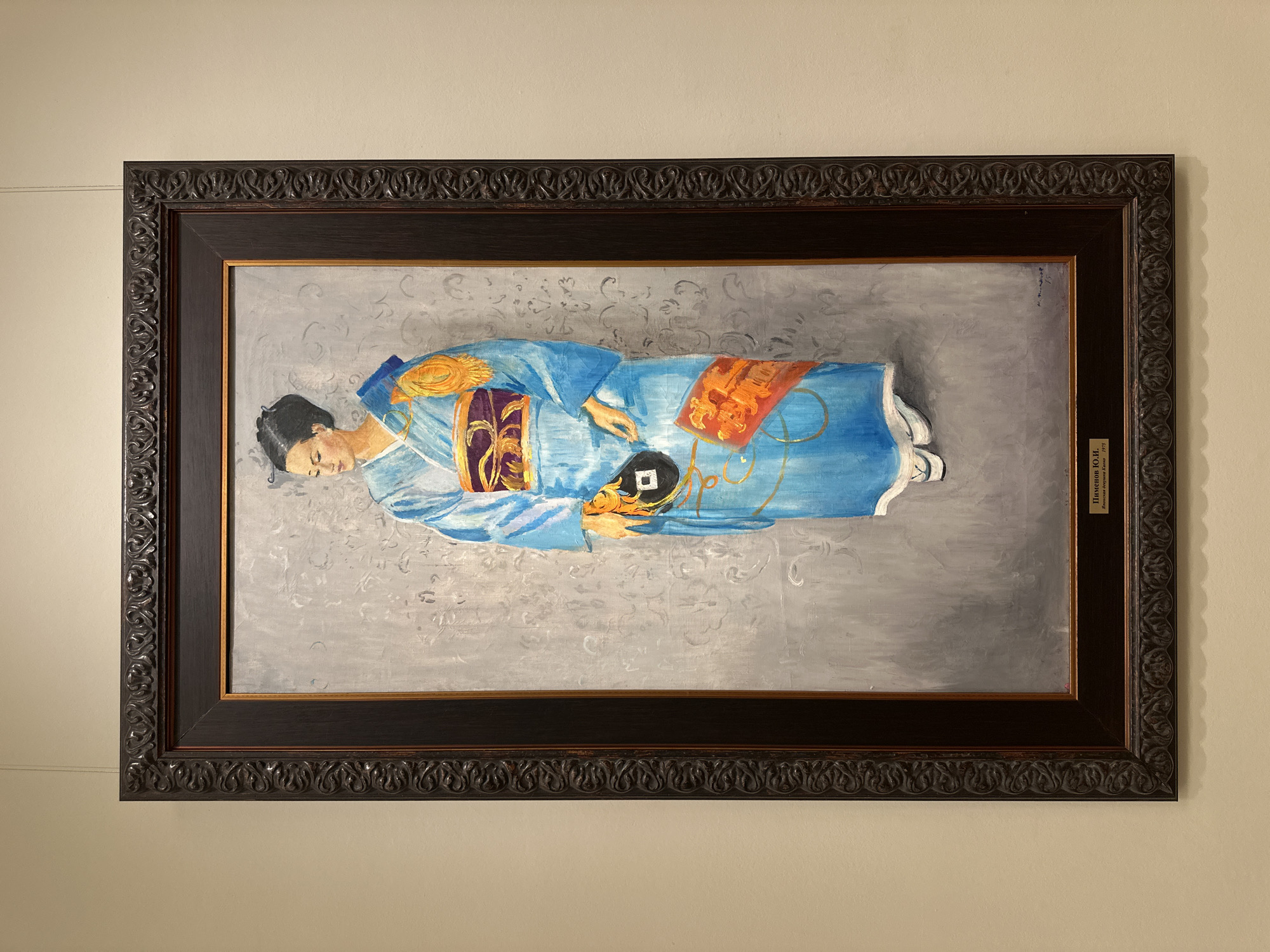 The image shows a framed painting hanging on a neutral-colored wall. The painting depicts a woman lying down, wearing a blue kimono with orange and purple accents. The background of the painting is a gray, patterned design. The frame is dark brown with ornate carvings. A small plaque is visible on the right side of the frame. The location is Unknown, Unknown.