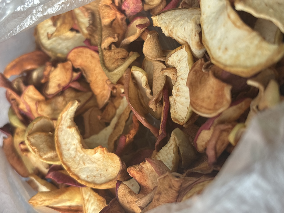 A close-up, overhead perspective captures a substantial quantity of irregularly shaped dried apple slices within a translucent plastic bag. The slices display a range of colors, from pale yellow and cream to reddish-brown and deep red, indicating the presence of apple skin on some pieces. The texture is shriveled and dry, characteristic of dehydrated fruit. The dried apple pieces are densely packed, occupying nearly the entire visual field. Edges of the clear plastic bag are visible along the upper left and right sides of the frame. No individuals, actions, or other discernible objects are present in the scene, which is located in Ivanovka, Russia.