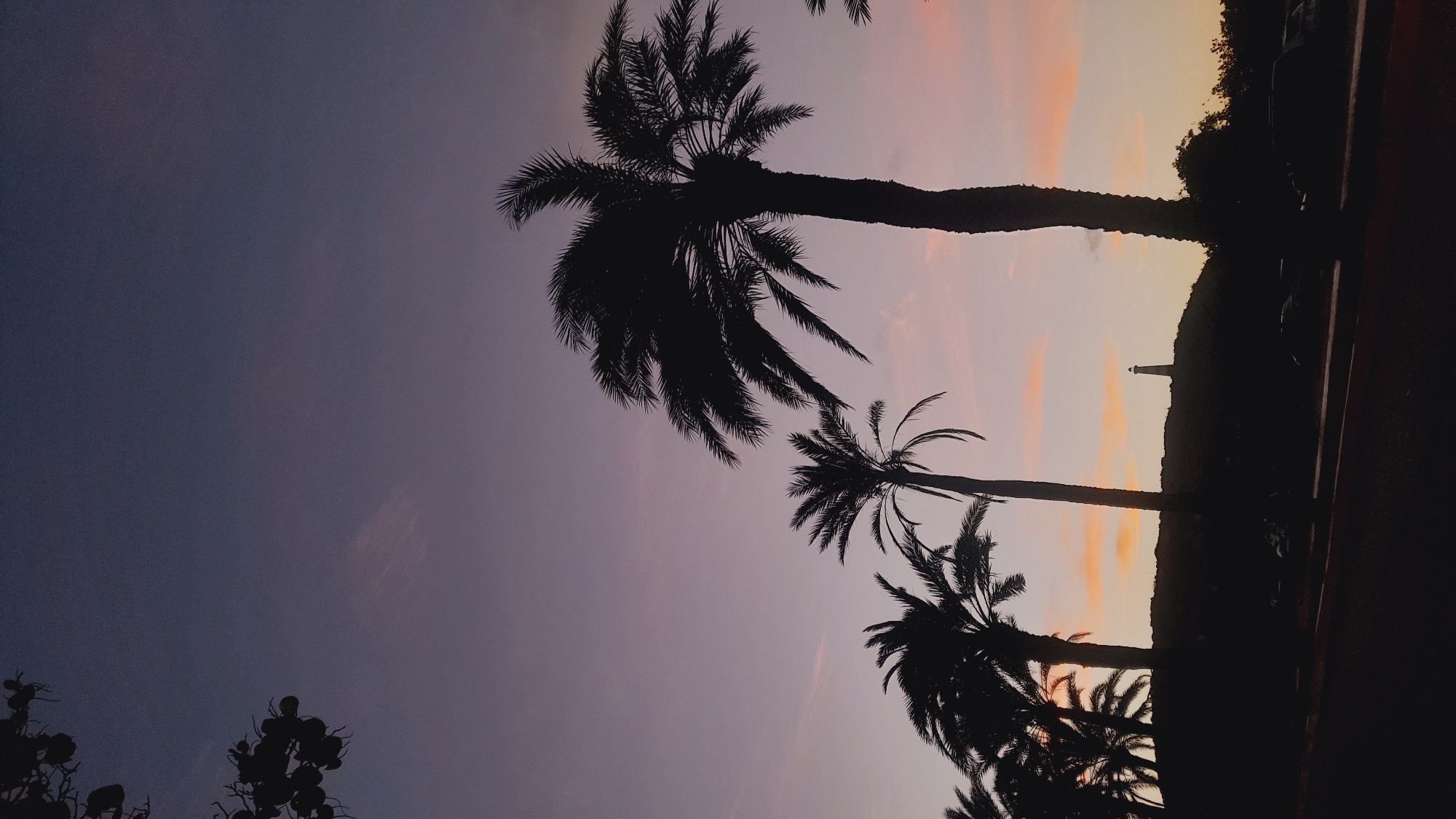 The image depicts a sunset scene with silhouetted palm trees in the foreground against a gradient sky of purple, pink, and orange. A lighthouse stands prominently on a hill in the background, overlooking a dark foreground which appears to be a road or parking area with a few vehicles. The overall atmosphere is serene and tropical, suggesting a coastal location. The image is captured from a low angle, emphasizing the height of the palm trees and the vastness of the sky. The scene is static, with no discernible human activity or interaction. The lighting suggests it is dusk or dawn, with the sun having recently set or about to rise.