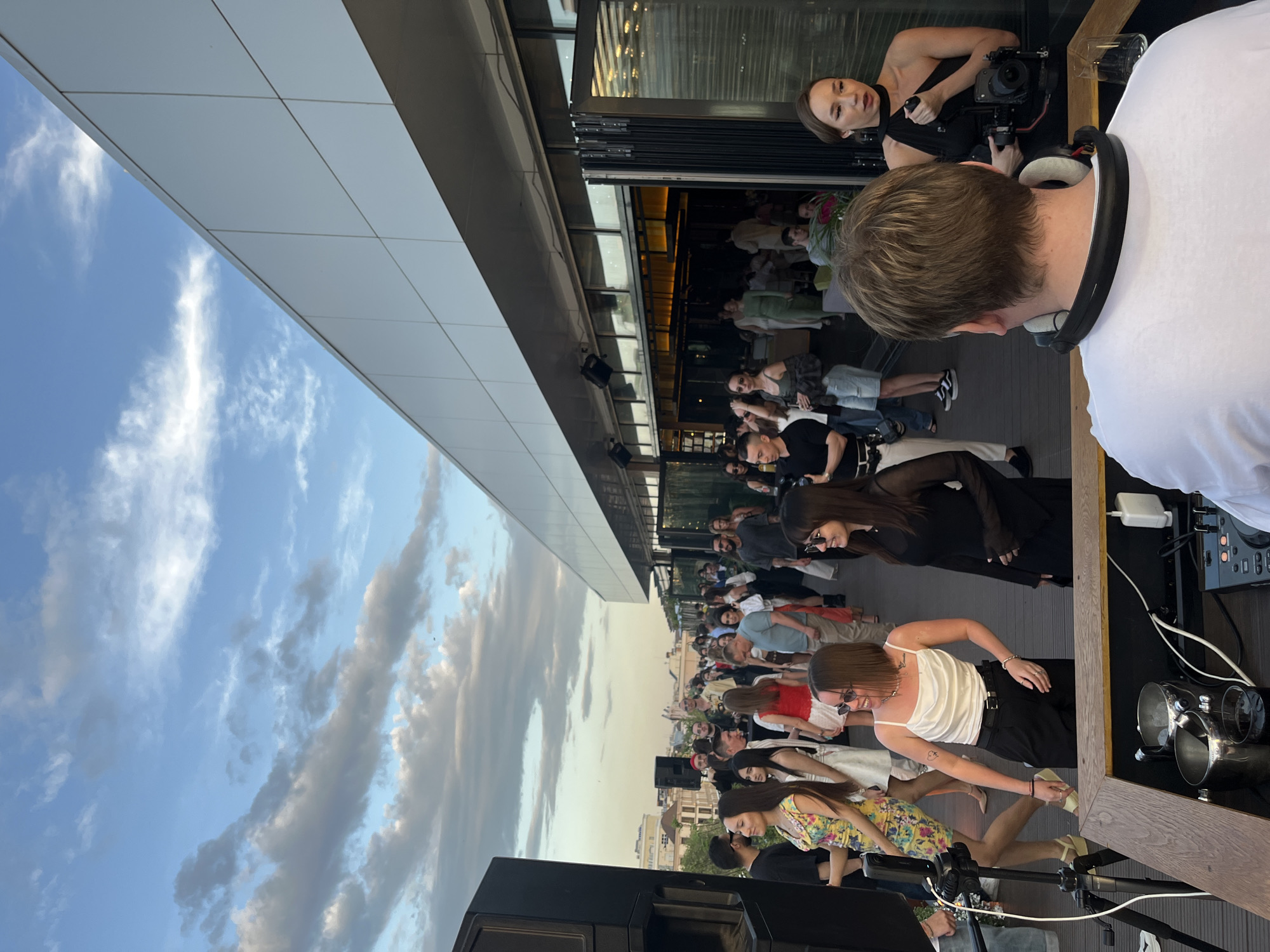 The image depicts an outdoor event on a rooftop venue in Kazan, Russia. A crowd of people is gathered, dressed in various casual and fashionable attire. The event appears to be social, with people standing and interacting. A DJ is present, seen from behind, with DJ equipment visible. A woman filming. In the background, buildings are visible on the horizon. The sky is visible and partly cloudy.