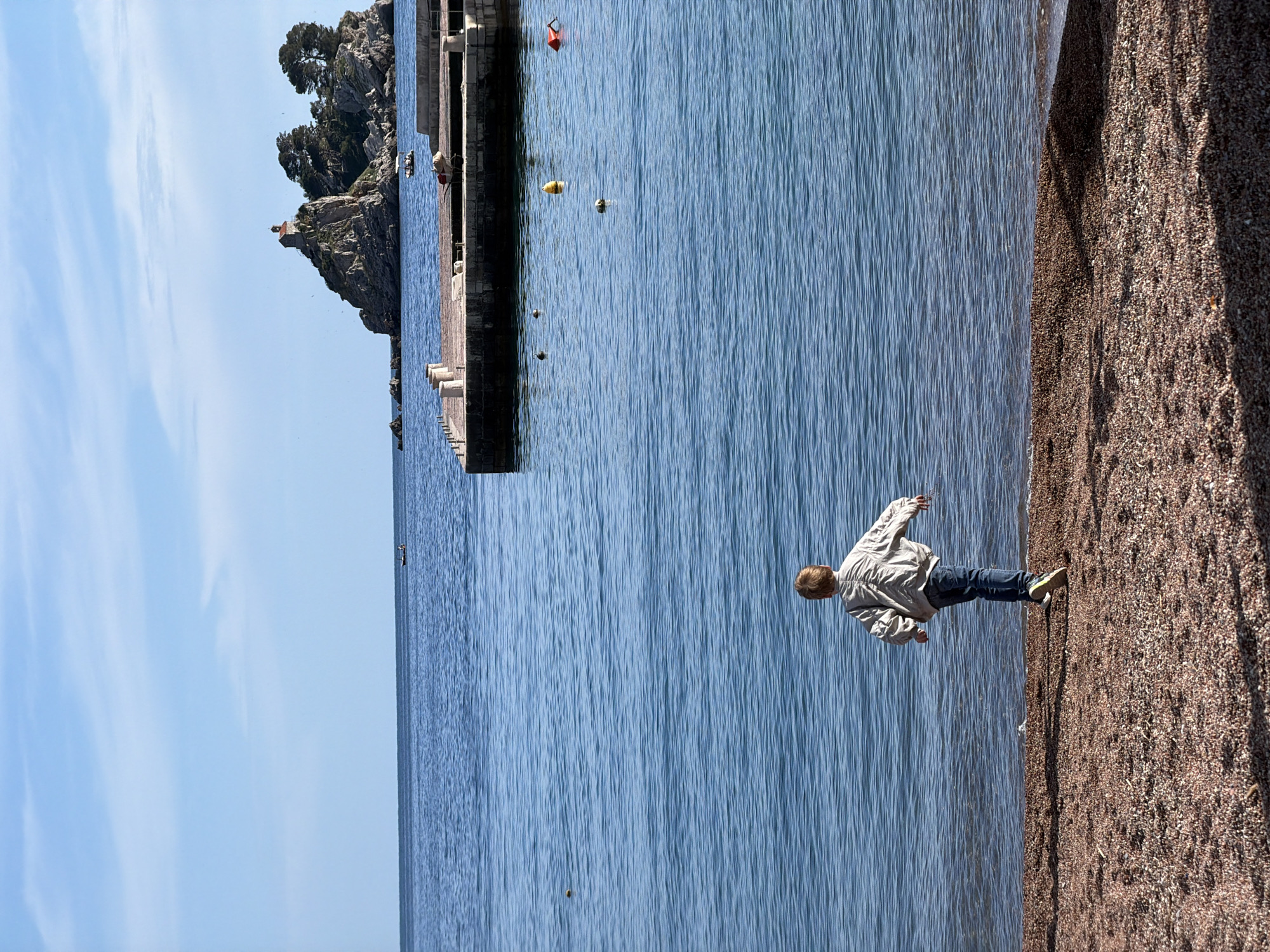 The image shows a person walking on a pebble beach alongside clear blue water on a sunny day. The location is Petrovac centar, Montenegro, indicated by the presence of the pier and the small island with a church on top, which is a known landmark of the city. The person is wearing a light jacket and jeans. The beach consists of small, reddish-brown pebbles. The pier extends into the water, and there are a few buoys visible in the distance.