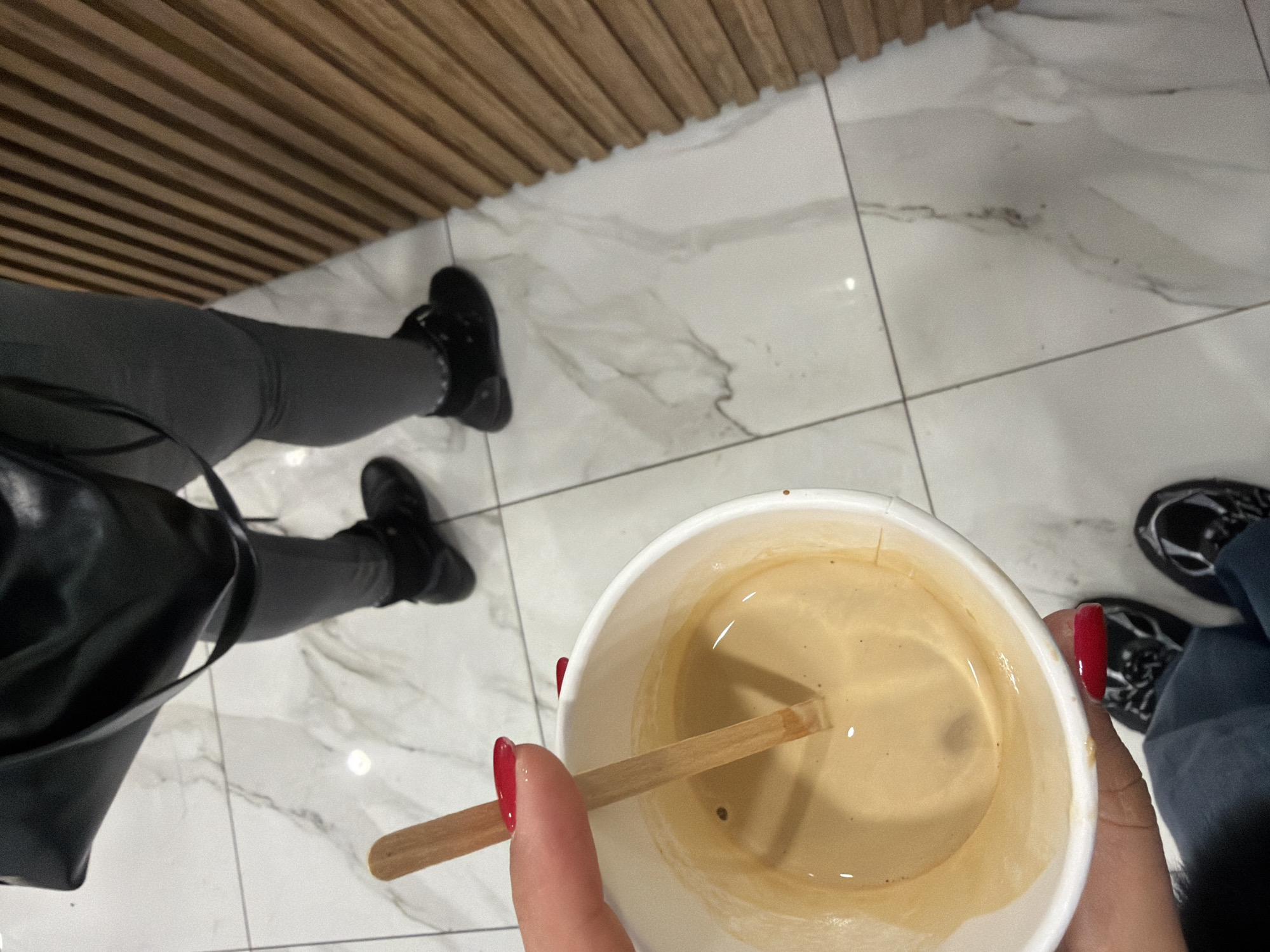The image shows an overhead view of a person stirring a cup of coffee with a wooden stick. The person is wearing gray leggings, black boots, and carrying a black bag. Another person is present, wearing black and white sneakers and blue jeans. The background consists of white marble tile flooring and a wooden slat wall. The location is potentially Sofia, Bulgaria.