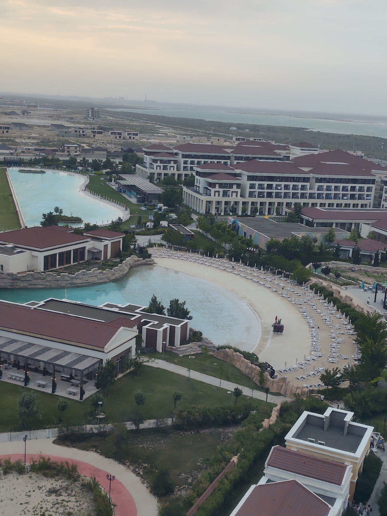 This is an aerial photograph of the Caspian Riviera complex in Aktau, Kazakhstan, taken on a cloudy day. The resort features a large artificial wave pool and a sandy beach area lined with lounge chairs and umbrellas. Several multi-story hotel buildings with red-tiled roofs are visible, as well as smaller structures that appear to be restaurants or service areas. In the distance, the Caspian Sea stretches to the horizon, with industrial facilities visible on the far shore. There are a few scattered individuals and a small group of people on a horse-drawn carriage on the beach. The overall impression is of a modern resort designed for leisure activities.