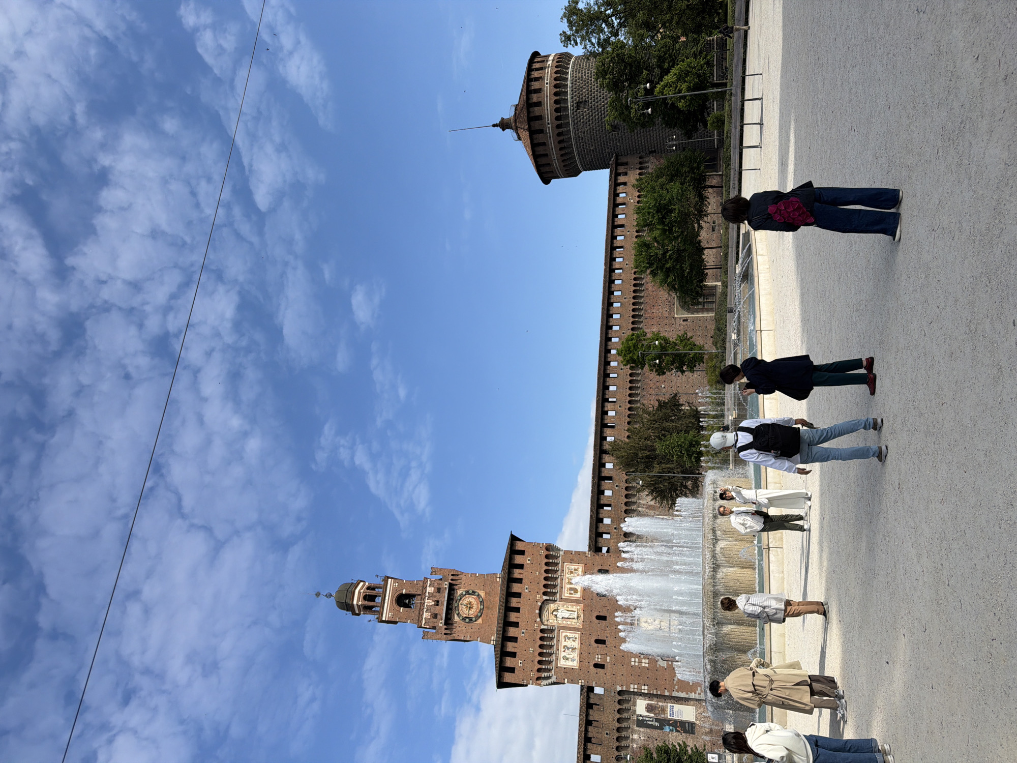 This outdoor image depicts the Sforza Castle in Milan, Italy, under a bright blue sky with scattered white clouds. The castle, constructed of reddish-brown brick, features prominent towers, battlements, and numerous windows. The central tower includes a clock face and a decorative top, while a cylindrical tower with a pointed, conical roof is visible on the right side of the castle complex.
In the foreground, a large, multi-tiered fountain sprays water, creating a lively atmosphere. Several people are scattered across the wide, light-colored paved ground in front of the castle and fountain. Most individuals are seen from behind or from a distance, suggesting casual movement or sightseeing. One person in dark clothing and blue jeans carries a reddish backpack. Another individual wears a white cap and a black backpack. People appear to be leisurely strolling or standing, observing the historical site.
The weather is clear and sunny, indicating daytime, likely late morning or early afternoon, given the bright light. Green trees are visible within and around the castle grounds. A thin black wire stretches across the upper part of the sky. On a lower section of the castle wall, a poster or banner is faintly visible, with the word "MILANO" somewhat legible, confirming the location. The scene captures a typical day at a major historical landmark, with tourists and locals enjoying the public space.