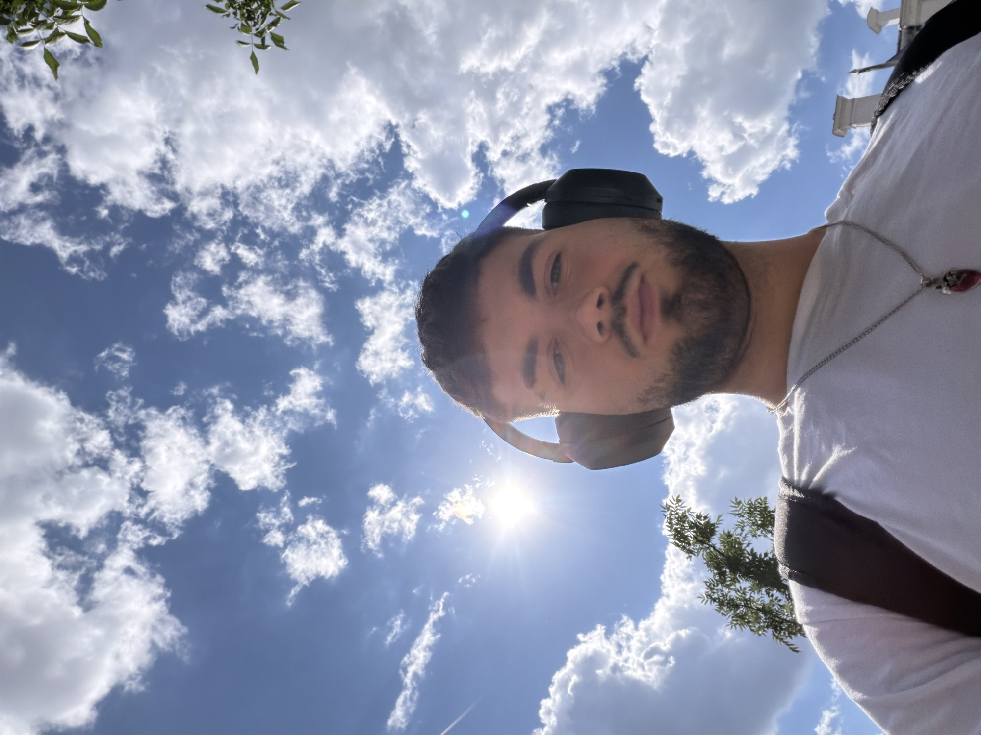 The media file is a close-up, low-angle selfie of a young man wearing black headphones and a white t-shirt. He has short, dark hair and a beard. A silver chain with a red pendant hangs around his neck. The man is looking up towards the sky.
The background is a bright blue sky filled with fluffy white clouds, with the sun prominently visible and creating lens flare. Green leaves from a tree are visible in the upper left corner, and part of a tree is also seen in the lower right. Some architectural elements, possibly part of a building with columns, are visible in the upper right.
The scene appears to be outdoors during the daytime, given the bright sunlight and clear sky. The overall mood seems relaxed and casual. The image captures a moment of enjoying the outdoors. There is no visible text in the media file.