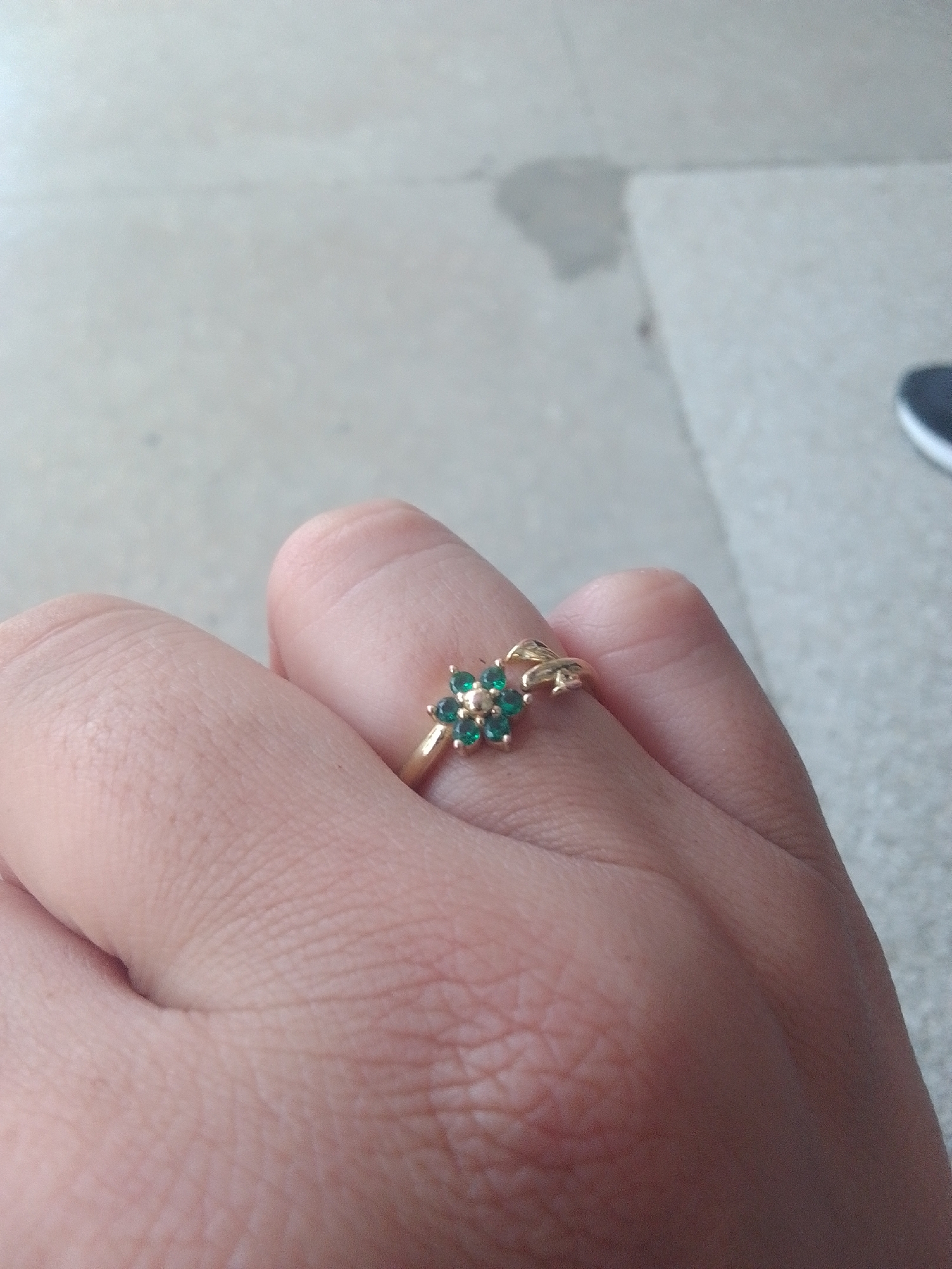 A close-up, eye-level shot shows a hand with a gold ring on the index finger. The ring features a flower design with green stones and a small, decorative element on the band. The background is a light-colored, textured surface, possibly a sidewalk or paved area. The lighting suggests it is daytime. The image is in focus on the ring and the finger, with the background slightly blurred.