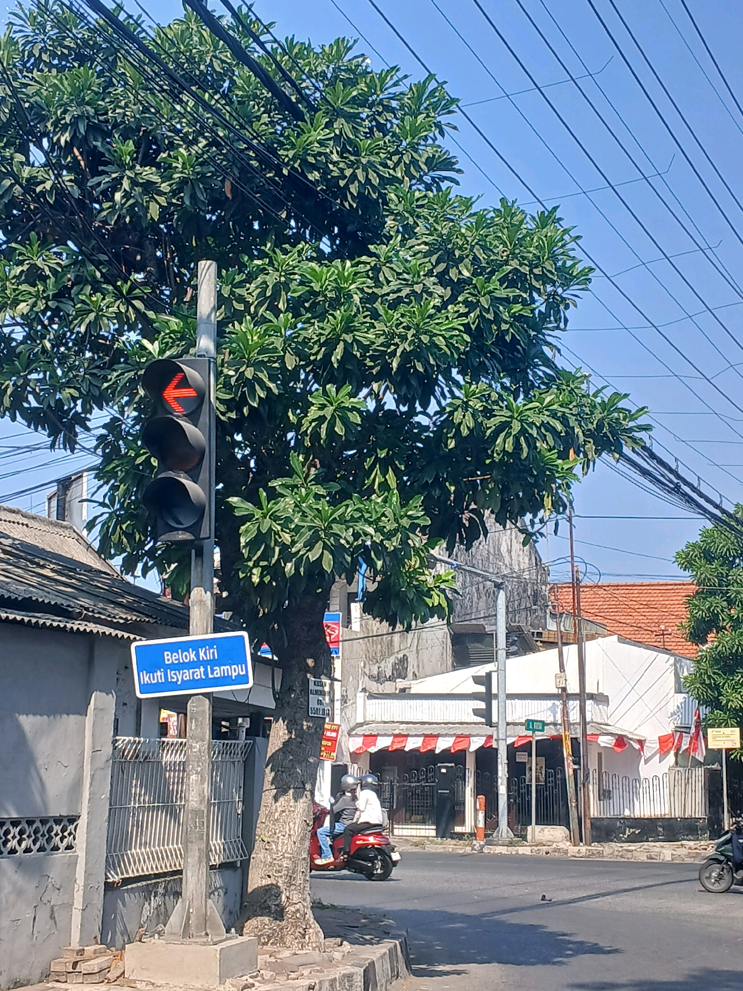 The image depicts a street scene in Surabaya, Indonesia, on a clear, sunny day. A traffic light shows a red left-pointing arrow, indicating that left turns are prohibited or restricted. A blue sign below the traffic light reads "Belok Kiri Ikuti Isyarat Lampu," which translates to "Left Turn Follow Traffic Signal."
In the foreground, a tree with dense green foliage partially obscures the traffic light and a building. A motorcycle carrying two people is visible turning to the left, potentially disregarding the traffic signal. To the left of the traffic light, a grey, textured wall and a white metal fence line the side of the road.
In the background, several buildings with tiled roofs and white facades are present. Red and white awnings or decorations adorn some of the buildings, suggesting a festive or nationalistic display. Power lines crisscross the sky, a common sight in urban Indonesian environments. Another motorcycle is seen in the distance on the right side of the image. The overall impression is a busy urban intersection where traffic rules might be loosely observed.