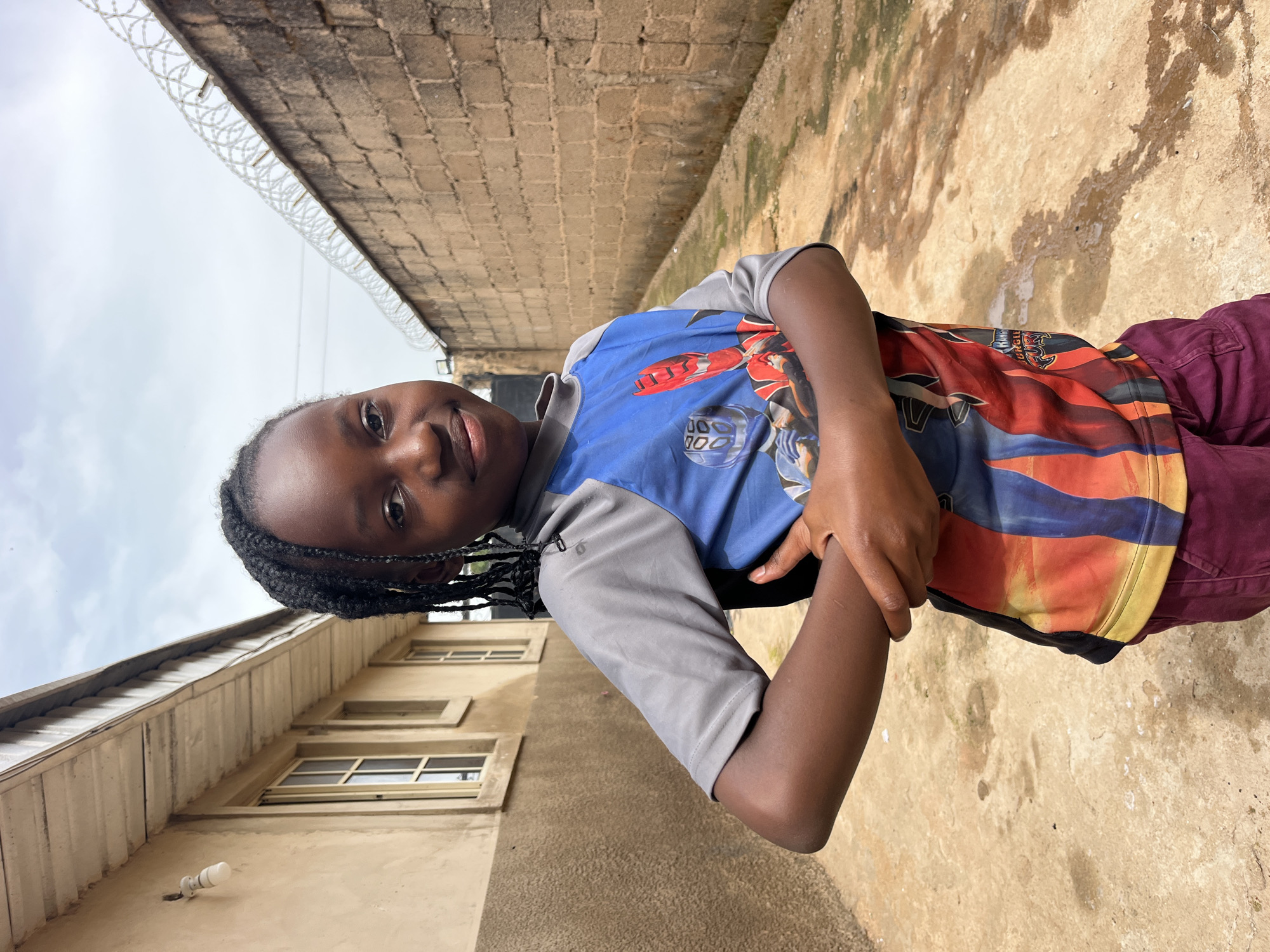 A young Black girl with braided hair stands outside, looking towards the camera with a slight smile. She is wearing a grey t-shirt with blue and red graphics of cartoon characters, and burgundy pants. Her arms are crossed in front of her. She is standing on a dirt path. To her left is a light-colored building with windows, and to her right is a brick wall topped with barbed wire. The sky is cloudy.