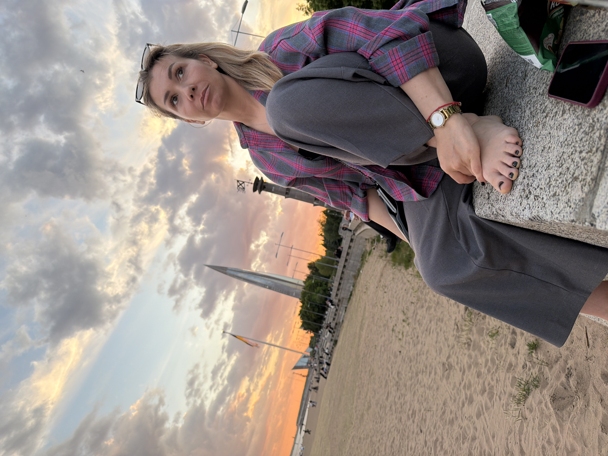 A woman with blonde hair and sunglasses perched on her head is seated on a concrete ledge, looking upwards with a pensive expression. She wears a grey and pink plaid shirt, grey wide-leg pants, a gold watch, and a red bracelet on her left wrist. Her bare feet, adorned with black nail polish, are visible, and she holds one of them with her hand. Next to her on the ledge are a green bag of "ХРУМ" (KHRUM) brand chips and a purple-cased smartphone.
The scene unfolds outdoors during the golden hour, possibly sunset, as warm orange and pink hues tint the clouds in the sky, contrasting with the lighter blue-grey clouds. The setting appears to be a riverbank or artificial beach, with a wide stretch of sand extending from the concrete ledge. In the distance, several streetlights line a promenade, and a prominent, tall, pointed monument – the Hero City Obelisk (Obelisk "To the Heroes of the City of Leningrad") – stands against the glowing sky. Other small figures of people can be seen along the promenade in the background. The location is consistent with an area in Saint Petersburg, Russia.