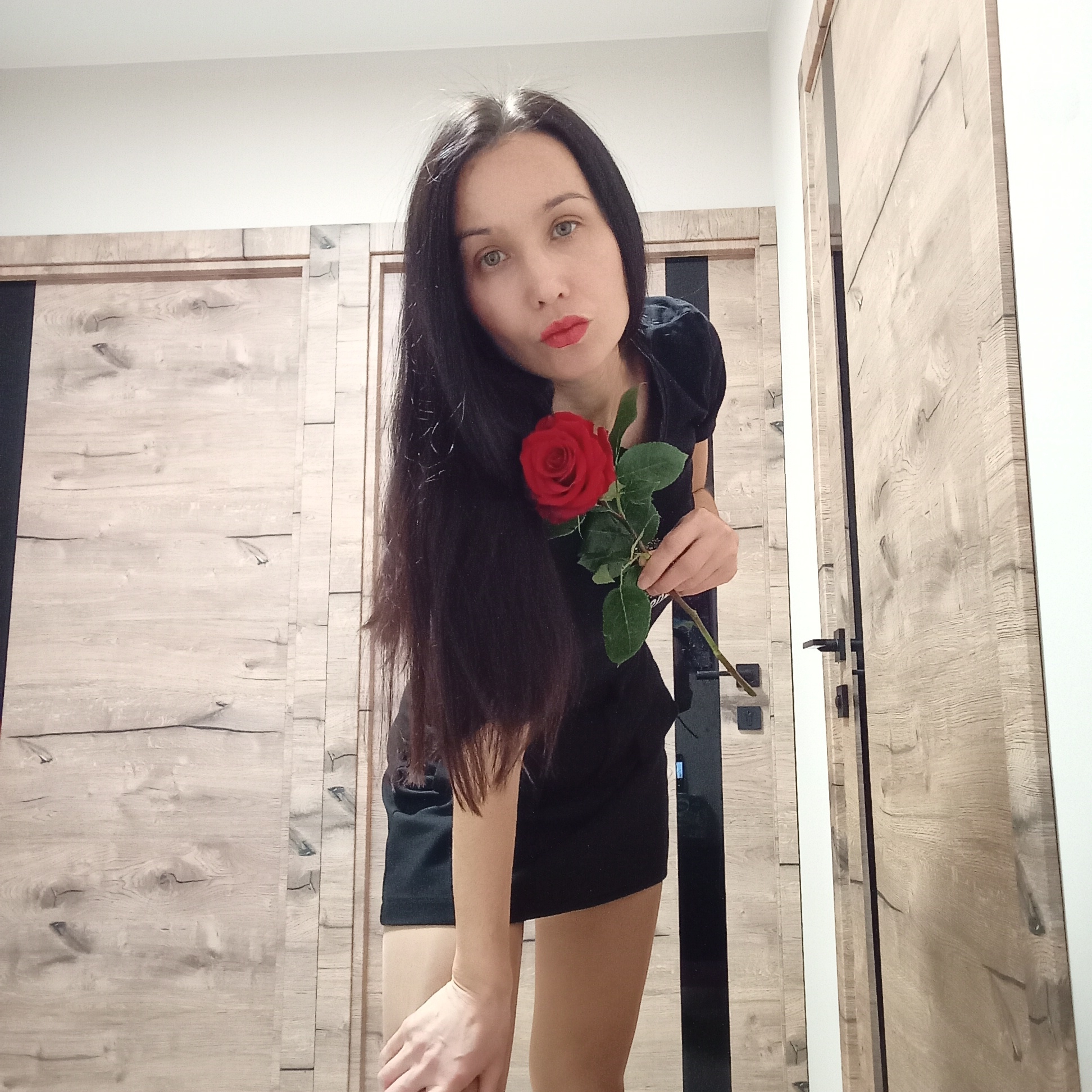 A woman is standing indoors holding a red rose. She has long, dark hair, fair skin, and red lipstick. She is wearing a black dress. The background consists of light wood-paneled doors. The location is in a room, possibly a hallway in the city of Pervomaysky, Russia. The photograph appears to be taken at close range.