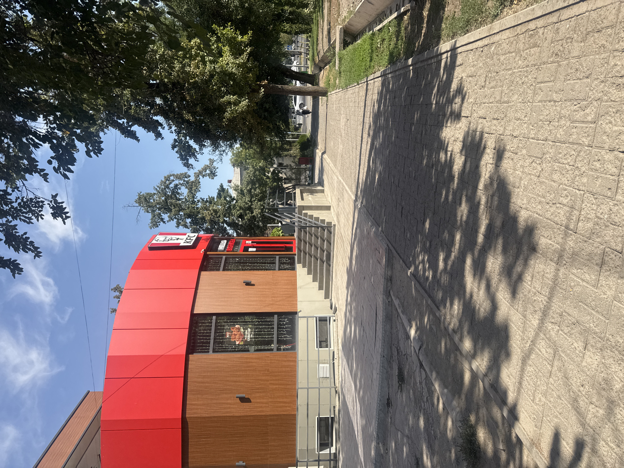 The image shows a KFC restaurant located in Bishkek, Kyrgyzstan. The building has a modern design with a bright red upper section and a brown wooden textured lower section. There are large windows displaying promotional posters, and a prominent KFC logo. A set of stairs leads up to the entrance, and a metal railing lines the side. A paved walkway extends from the building into the distance, bordered by a grassy area with a drainage channel. Trees with green foliage line the street, casting shadows on the walkway. In the background, a few cars and people are visible, indicating a typical urban environment. The sky is blue with some scattered clouds.