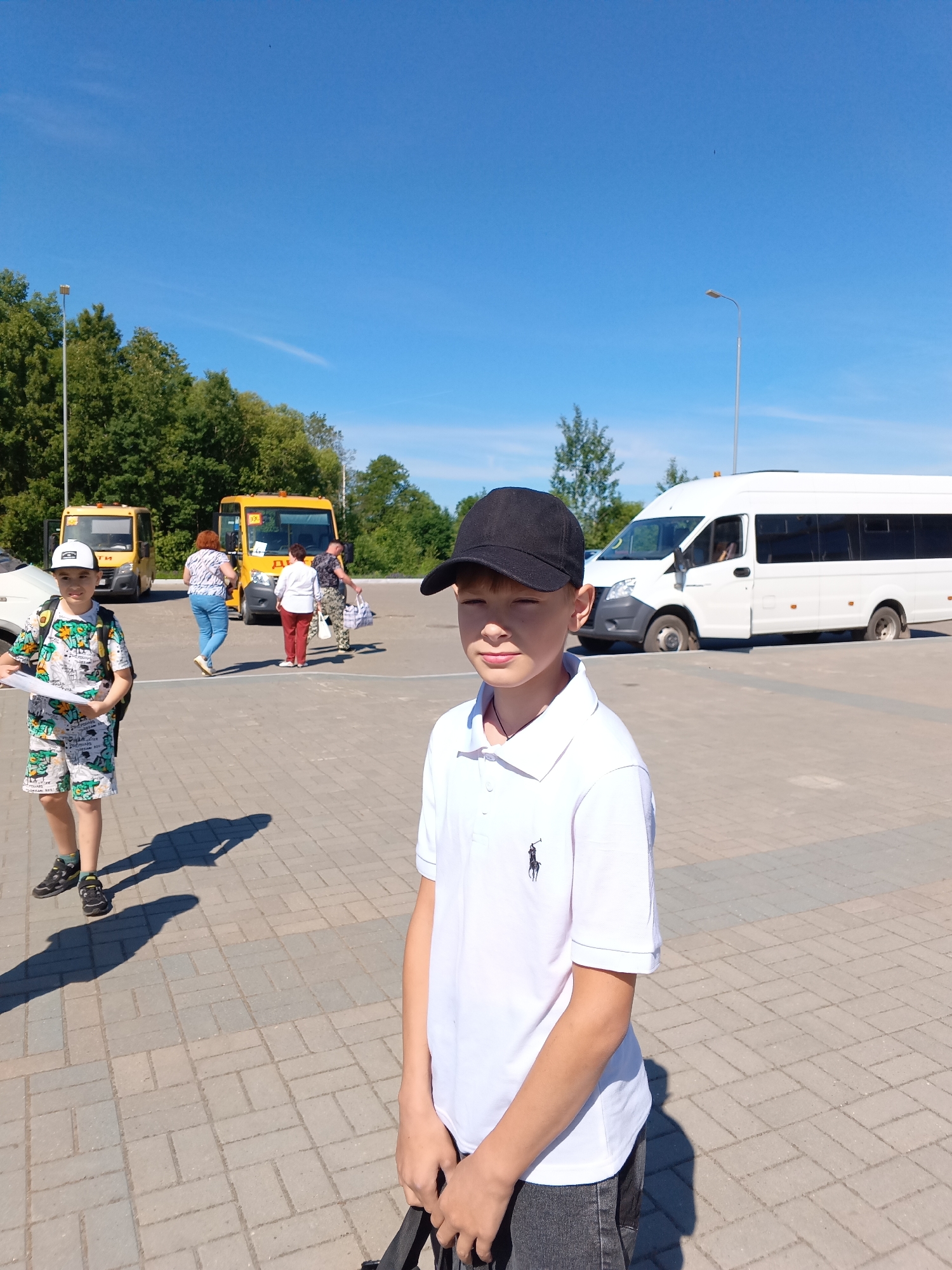 The image depicts a bright, sunny outdoor scene in Sudogda, Russia, likely a paved parking area or square, under a clear blue sky. The ground is paved with rectangular and square interlocking pavers in shades of grey and tan, arranged in a herringbone pattern, showing distinct shadows cast by the sun.
In the foreground, slightly to the right, stands a young boy, approximately 8-12 years old. He has light skin, wears a black baseball cap, a white polo shirt with a dark embroidered emblem on the left chest (resembling a Polo Ralph Lauren logo), and dark grey jeans. His eyes are partially closed, possibly squinting against the bright sunlight, and his hands are clasped in front of him, holding a dark object or strap.
In the mid-ground, to the left, another young boy of similar age is walking towards the left side of the frame. He wears a white baseball cap, a white t-shirt and matching shorts with a multi-colored (green, white, black) pattern, and dark sneakers. He carries a dark backpack and holds several white papers in his left hand. His shadow is visible on the pavement to his right.
In the background, several individuals and vehicles are present. Two yellow minibuses are parked on the left, both bearing Cyrillic text, including "ШКОЛА" (SCHOOL), indicating they are school buses. Near these buses, at least three adults are visible: one woman with reddish-brown hair in a white patterned top and blue jeans, walking towards the buses with a white bag; another woman in a white top and red pants, standing and carrying a light-colored bag; and a man in a camouflage-patterned shirt, standing near the second yellow bus with two bags.
To the right of the background, a white minibus or large van is parked, with its passenger side door appearing to be open or ajar. This vehicle has dark tinted windows in the rear section.
The background also features a dense line of green deciduous trees, and at least two tall, slender lampposts with modern light fixtures rise above the trees and vehicles. The sky is a vibrant blue with a few faint wispy clouds and a small, dark, unidentifiable speck visible in the upper central portion. White line markings are visible on the pavement near the white van.