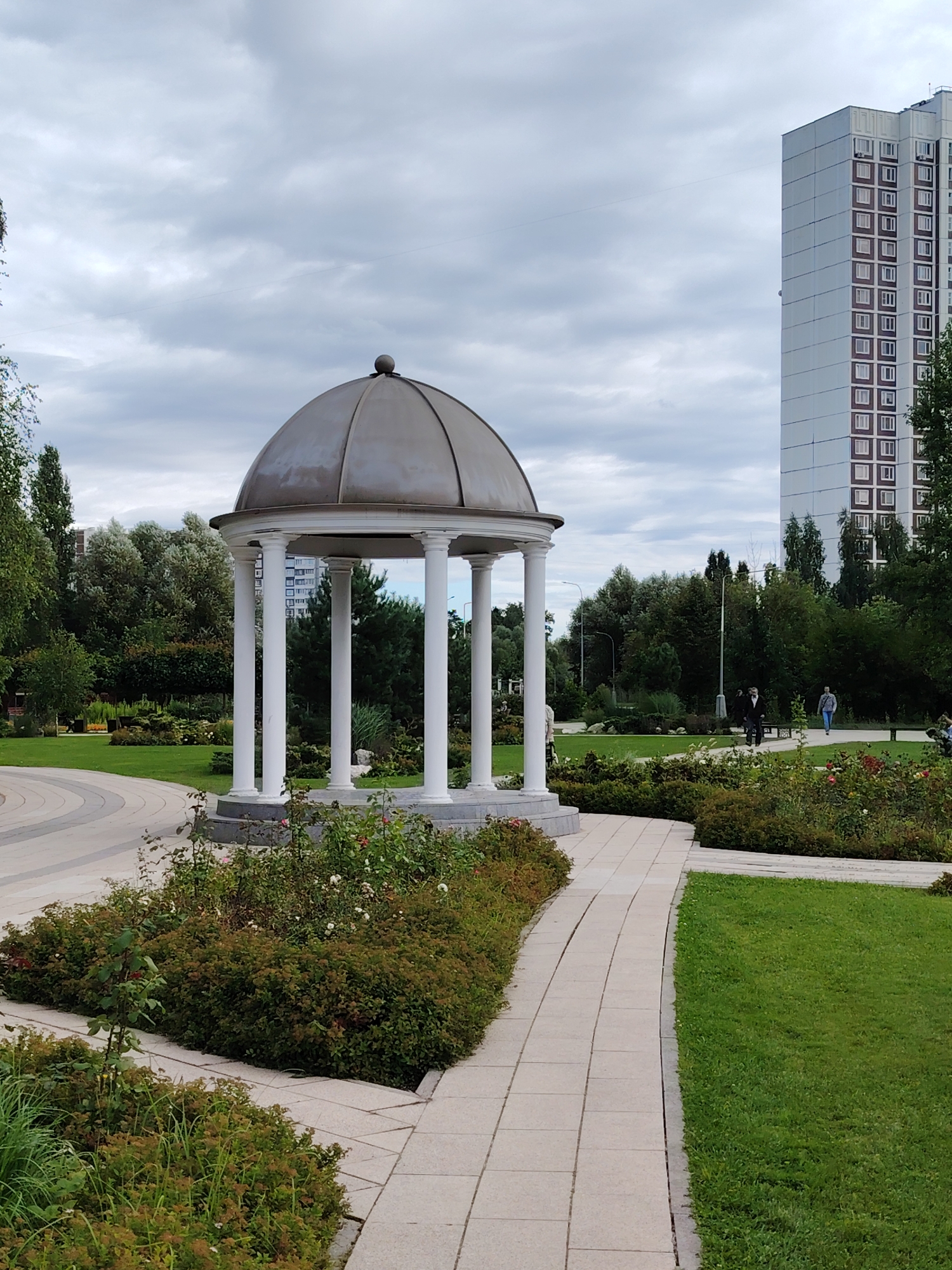 The image depicts a park scene in Zelenograd, Russia, featuring a white-columned gazebo with a domed roof in the foreground. The gazebo is surrounded by greenery, including rose bushes and manicured shrubs. A light-colored paved walkway curves through the park, bordered by green grass on one side and dense vegetation on the other. In the background, a tall, modern residential building with numerous windows stands against a cloudy sky. Several people are visible walking along the paths in the distance, suggesting a public space. The overall environment appears to be a well-maintained urban park.
