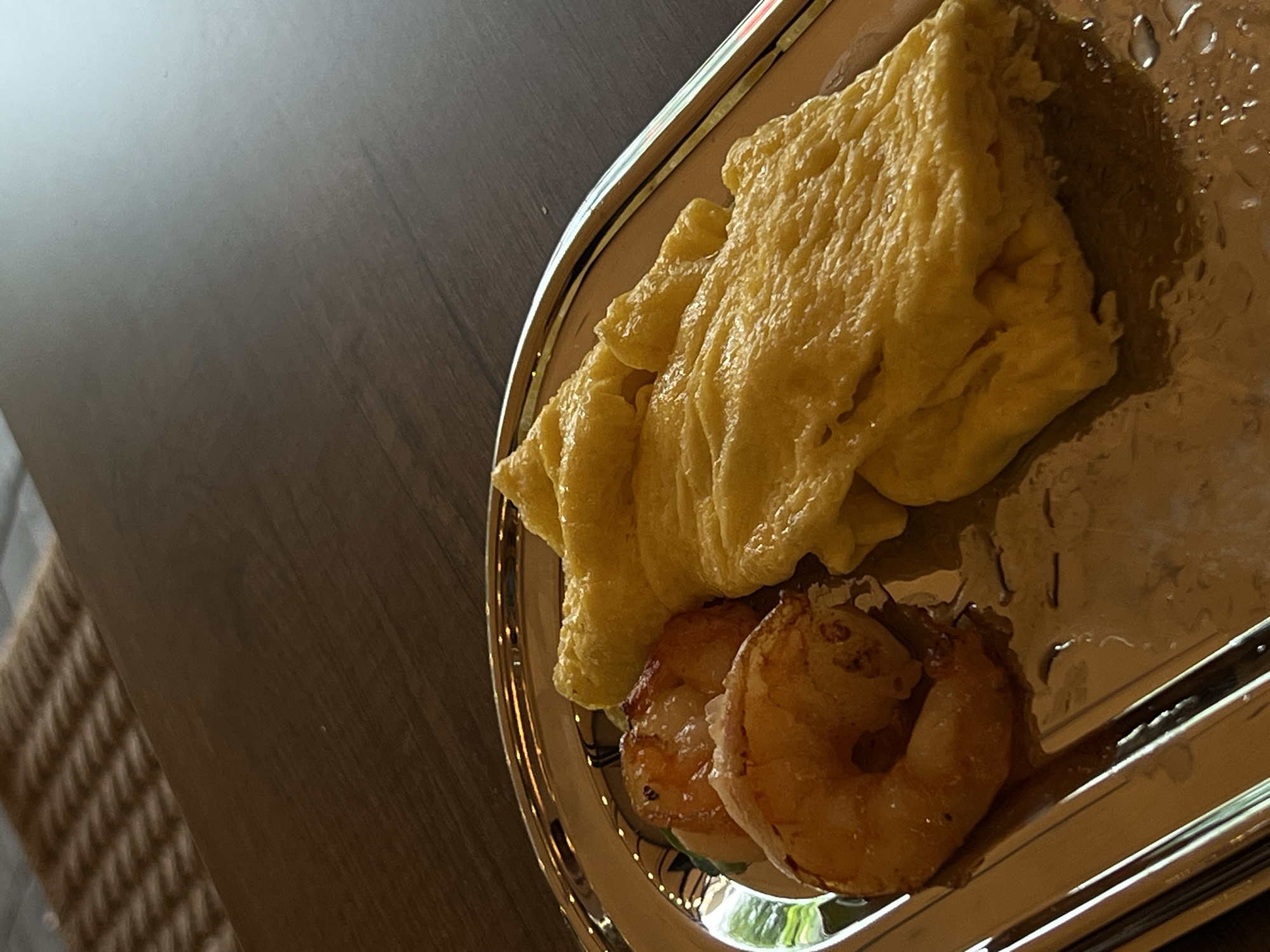 The image depicts a plate with an omelet and two fried shrimp, likely photographed in a restaurant in Yerevan, Armenia. The plate appears to be metallic with a textured surface and a decorative rim. A fork rests on the plate's edge. The table's surface is dark wood, and a wicker texture is visible on the left, possibly part of a chair. No people are directly visible. The focus is on the food and the arrangement of items on the plate.