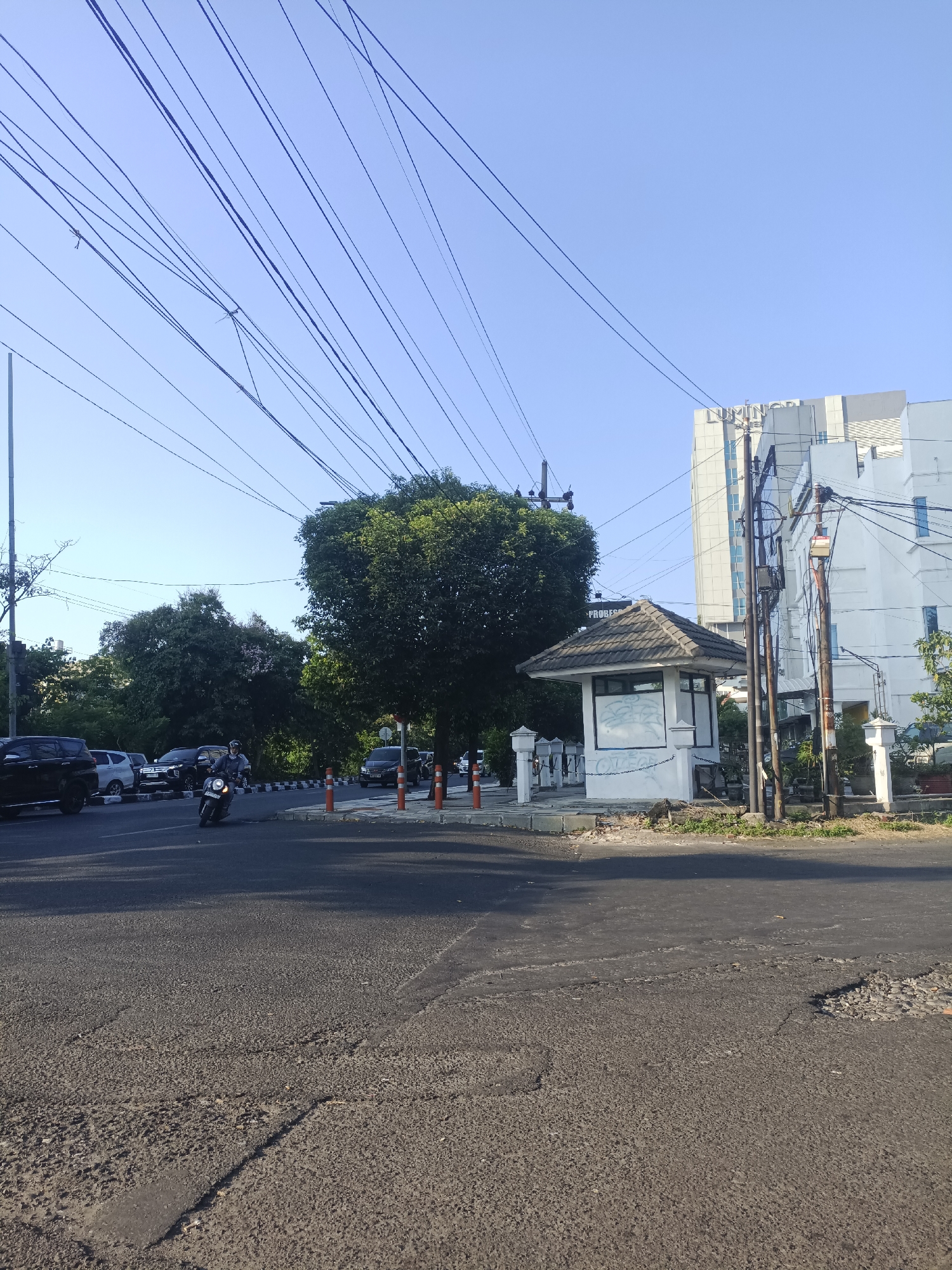 The image depicts a street scene in Surabaya, Indonesia, characterized by a clear blue sky and overhead electrical wires. A modern, multi-story building, possibly a hotel or office complex, with a sign reading "LUMINOR" is visible in the background. In the foreground, a paved road shows signs of wear and tear, with potholes and patches. A small, grey, open-sided kiosk with a tiled roof stands on the sidewalk, its walls covered in graffiti. Orange traffic cones are placed along the curb. Several vehicles are present, including a black SUV, a white car, and a motorcycle with a rider wearing a helmet and a grey jacket, traveling on the road. Trees with dense green foliage line the street, contributing to the urban landscape.