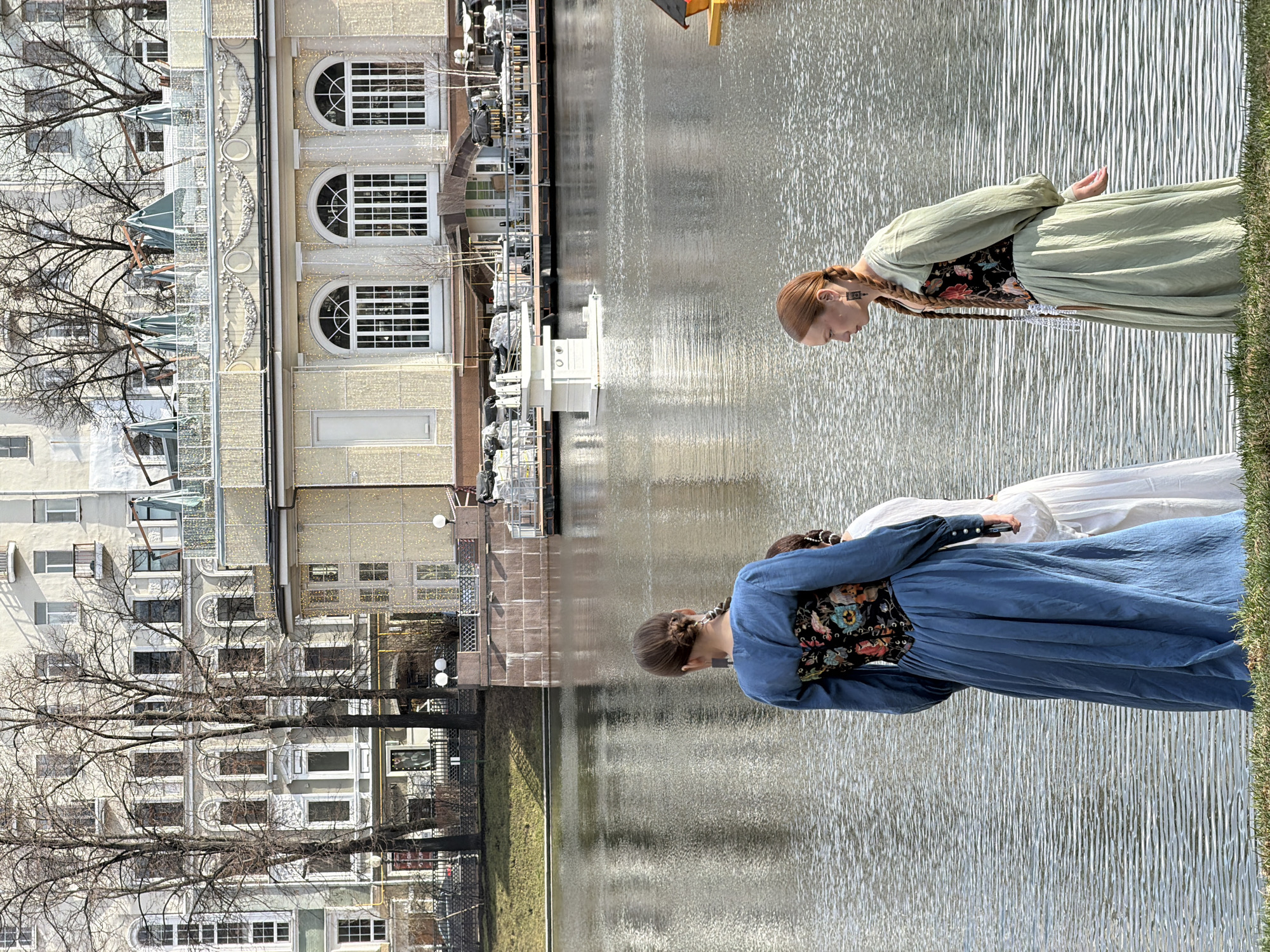 The image depicts a street scene in Moscow, Russia, alongside a body of water (likely a canal or the Moscow River). Two women in period-style dresses stand near the water's edge. One wears a blue dress with a floral-patterned bodice, and the other is in a light green dress, also with a dark floral bodice. The woman in the green dress has long red hair braided down her back. Both appear to be looking down towards the water.
Behind the women, the cityscape is visible. There are a series of buildings, some with ornate architectural details, including arched windows. Bare tree branches extend into the frame, suggesting it might be early spring or late fall. There are some modern structures and what appears to be construction or renovation work near the water. The body of water reflects the buildings and the sky.