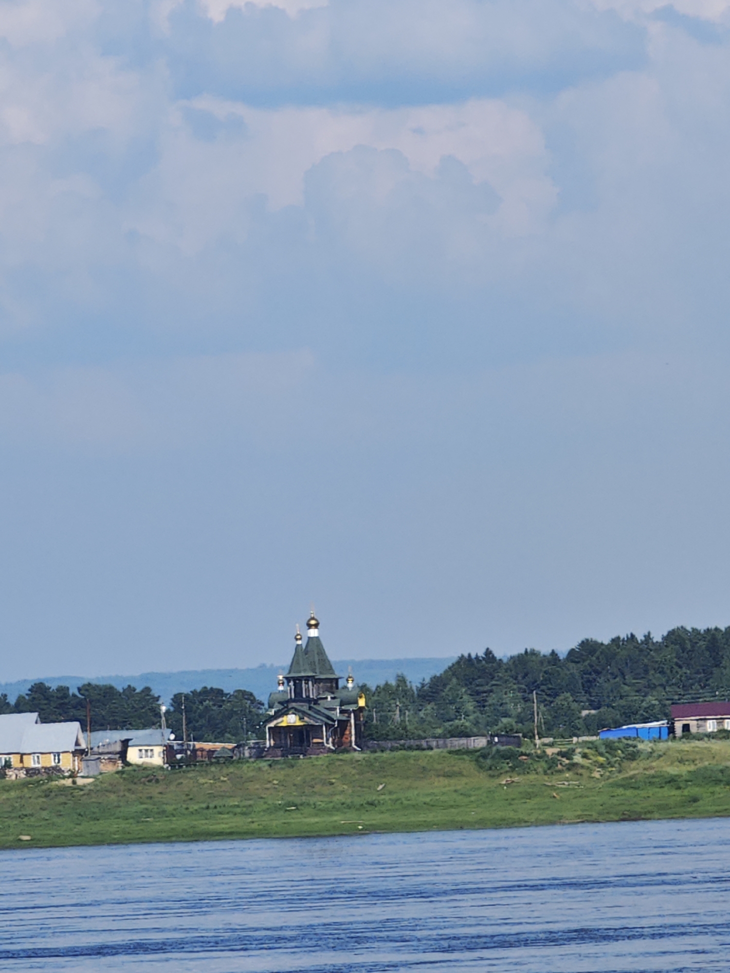 The image shows a body of water in the foreground, with ripples on the surface. In the middle ground, a green, grassy bank rises, leading up to a settlement. Several buildings are visible, including a distinctive wooden church with green roofs and golden domes, and several houses. Behind the settlement, a dense forest stretches across the horizon, with rolling hills in the far distance. The sky above is a pale blue with scattered white clouds. The scene is tranquil and appears to be a rural setting.