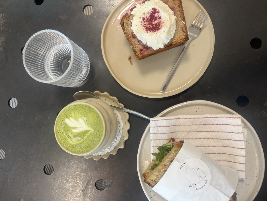 The scene depicts a tabletop arrangement of food and beverages at KLAY Keramik & Café, located at Eisenbahnstraße 19, 67655 Kaiserslautern, Germany. The dark, textured table surface features a variety of items. In the upper center, a beige ceramic mug with a ridged exterior contains a bright green matcha latte, distinguished by a white leaf-patterned latte art. The mug rests on a matching ruffled ceramic saucer, with a white doily underneath. A silver spoon is positioned on the saucer. To the upper right, a clear, vertically ribbed glass tumbler holds a clear liquid. On the lower left, a round, speckled light-colored ceramic plate presents a sandwich, partially wrapped in white paper bearing the "KLAY Keramik & Café" logo. Visible sandwich components include whole-grain bread and green leafy vegetables. A folded white napkin with thin red stripes is placed beneath the sandwich wrap. On the lower right, an irregular beige ceramic plate holds a slice of cake or loaf, exhibiting red fruit pieces within its light-brown structure. This item is topped with a substantial dollop of white cream, sprinkled with red powder. A silver fork rests diagonally across this plate. No individuals are visible in the frame.